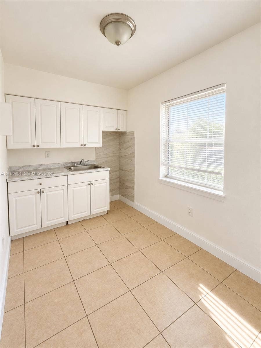 White shaker-style cabinetry, neutral stone-patterned countertops, stainless steel sink, large-format ceramic tile flooring, and a flush-mount ceiling light fixture