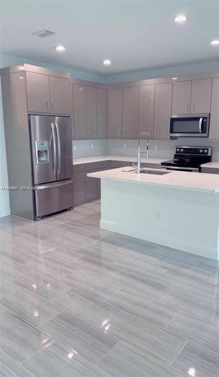 Kitchen featuring high-gloss cabinetry, stone countertops, stainless steel appliances, a center island with an integrated sink, and large-format porcelain tile flooring