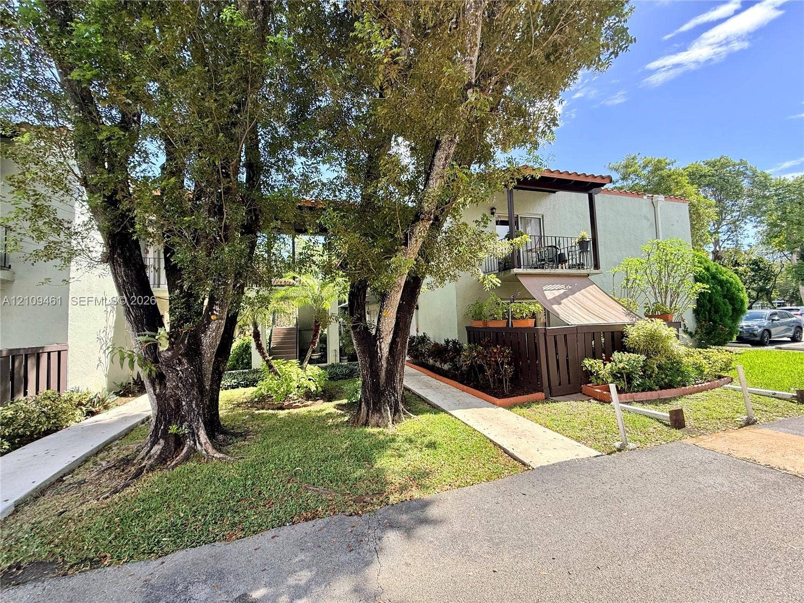 10776 N Kendall Drive, #F11, Miami, FL 33176 Two-story exterior featuring light-colored stucco siding, a tiled roofline, second-level balcony with railings, ground-level patio with privacy fencing, and mature trees