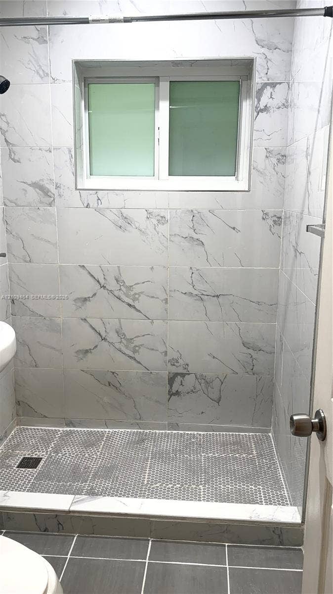 Walk-in shower enclosure featuring large-format veined tile surround, hex-pattern mosaic floor tiling, integrated horizontal window, and dark-finish square shower drain