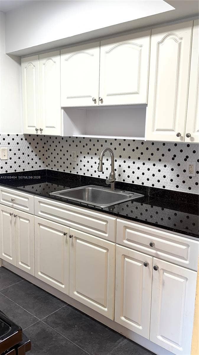 White shaker-style cabinetry with recessed panels, dark stone countertops, stainless steel undermount sink, gooseneck faucet, and geometric-patterned backsplash