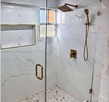 Walk-in shower featuring white large-format tile walls, mosaic stone tile flooring, a built-in shower niche, frameless glass enclosure, and metallic gold-finish fixtures including a rainfall showerhead and handheld sprayer