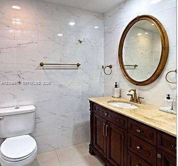 Bathroom vanity featuring a natural stone countertop, undermount sink, dark wood-finish cabinetry, and an oval mirror, complemented by floor-to-ceiling wall tiling and gold-toned fixtures