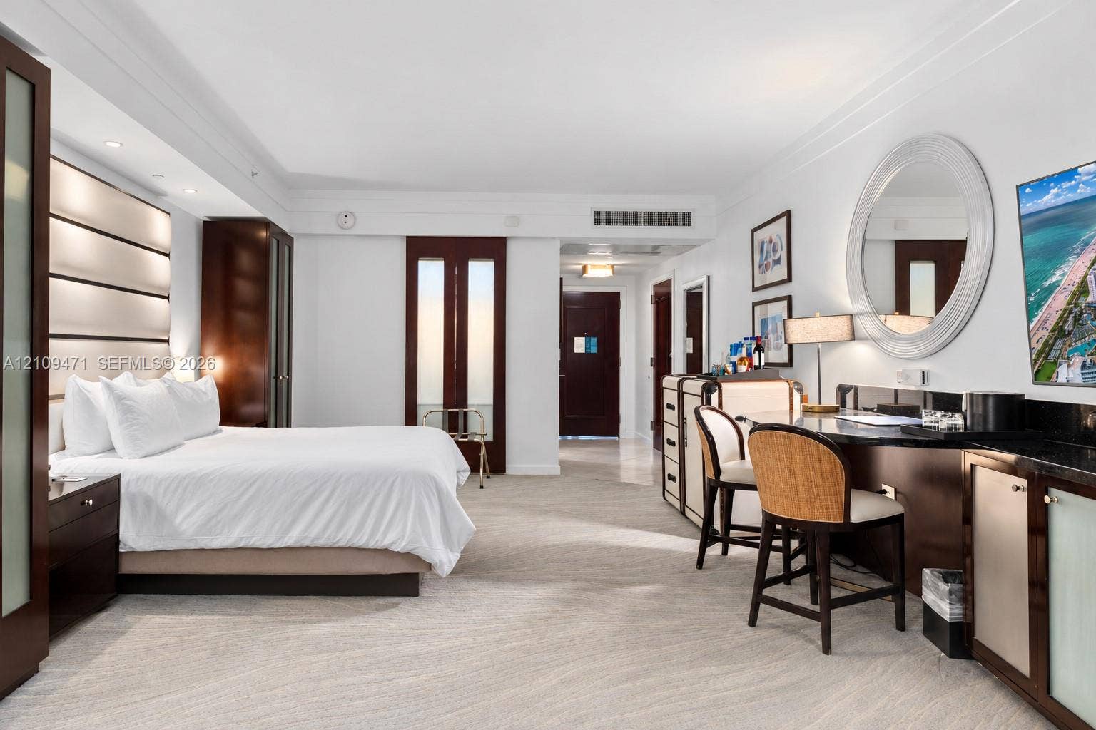 Spacious room featuring a wall-mounted panel headboard, dark wood-finish cabinetry, a built-in stone countertop desk, a circular wall mirror, and neutral-toned carpeting
