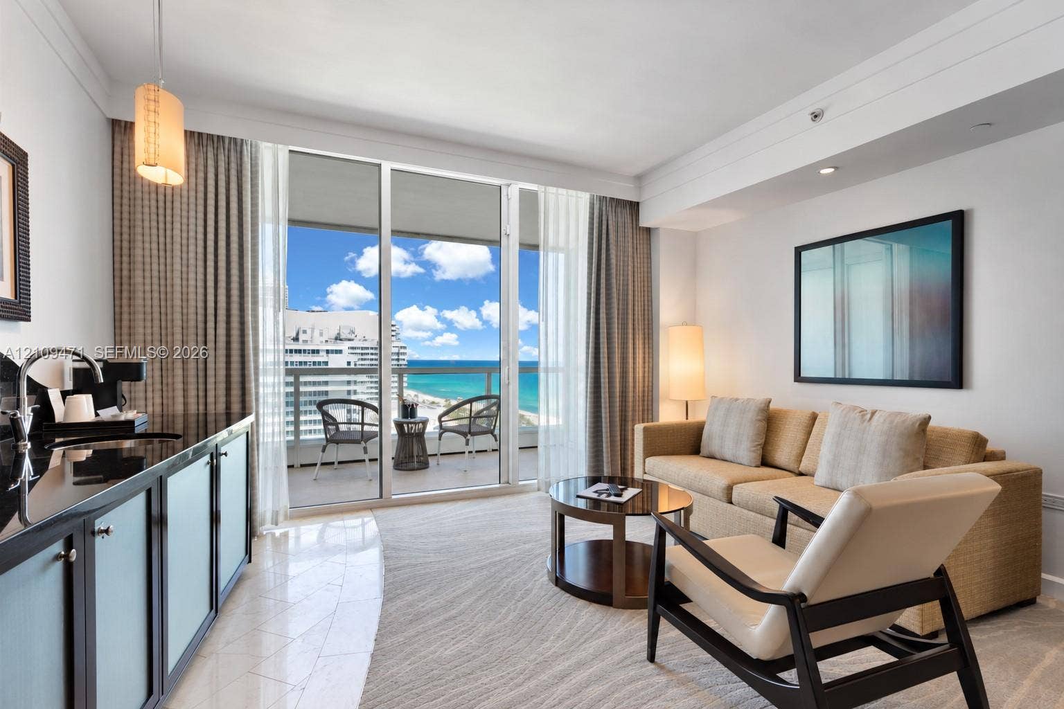 Living area featuring floor-to-ceiling glass sliding doors, private balcony with ocean frontage, wet bar with dark stone countertop, and polished tile flooring