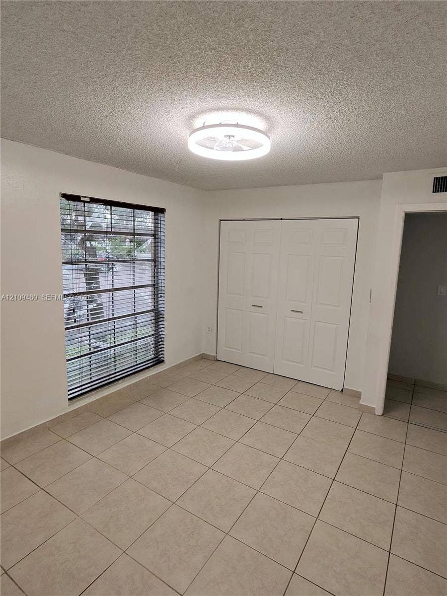 Neutral tile flooring, circular flush-mount LED ceiling fixture, dual-panel closet doors, and a window with horizontal blinds