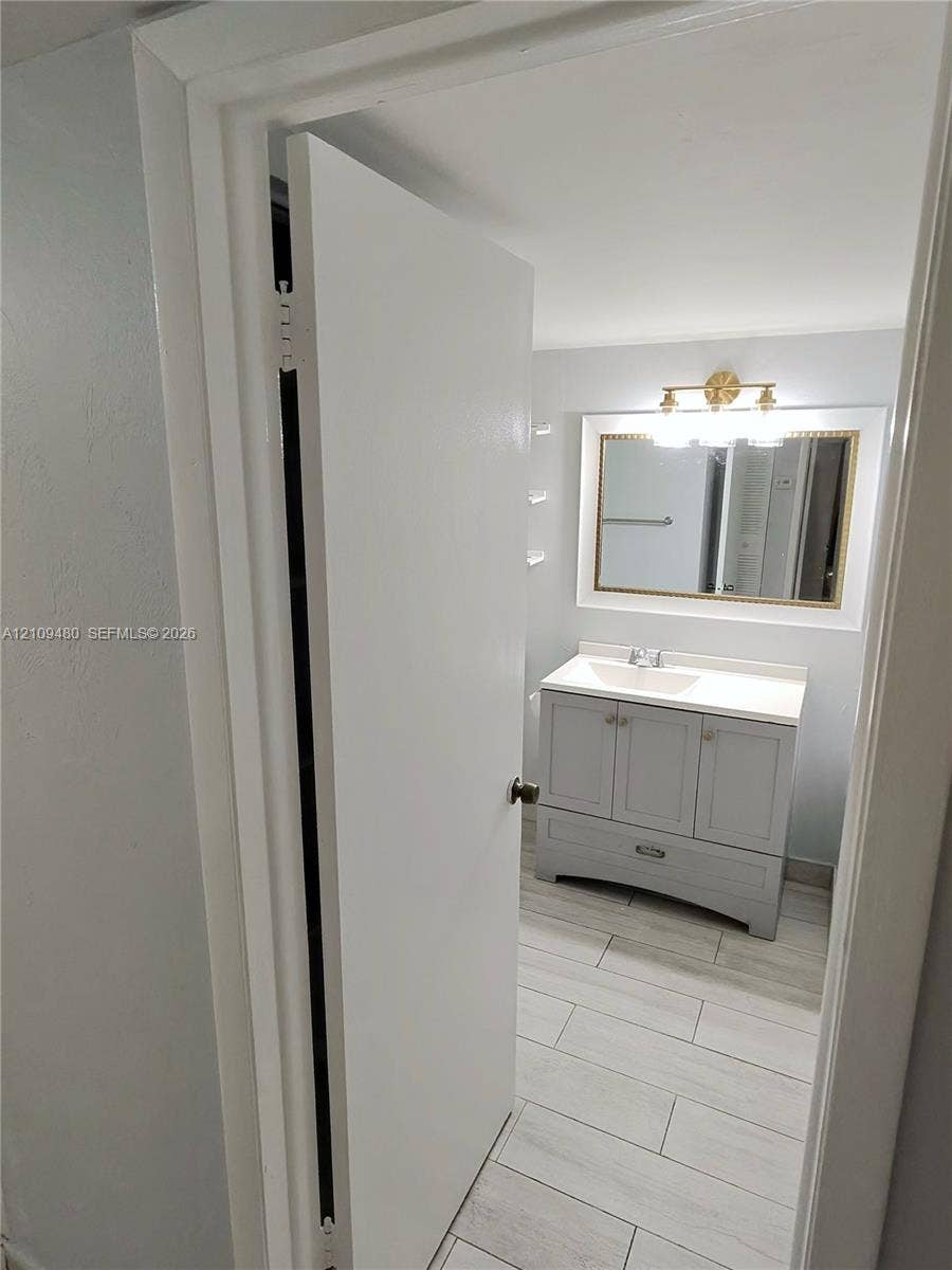 Bathroom featuring a grey vanity with a rectangular sink, stone-finish countertop, framed wall mirror, multi-bulb vanity light, and light-toned tile flooring