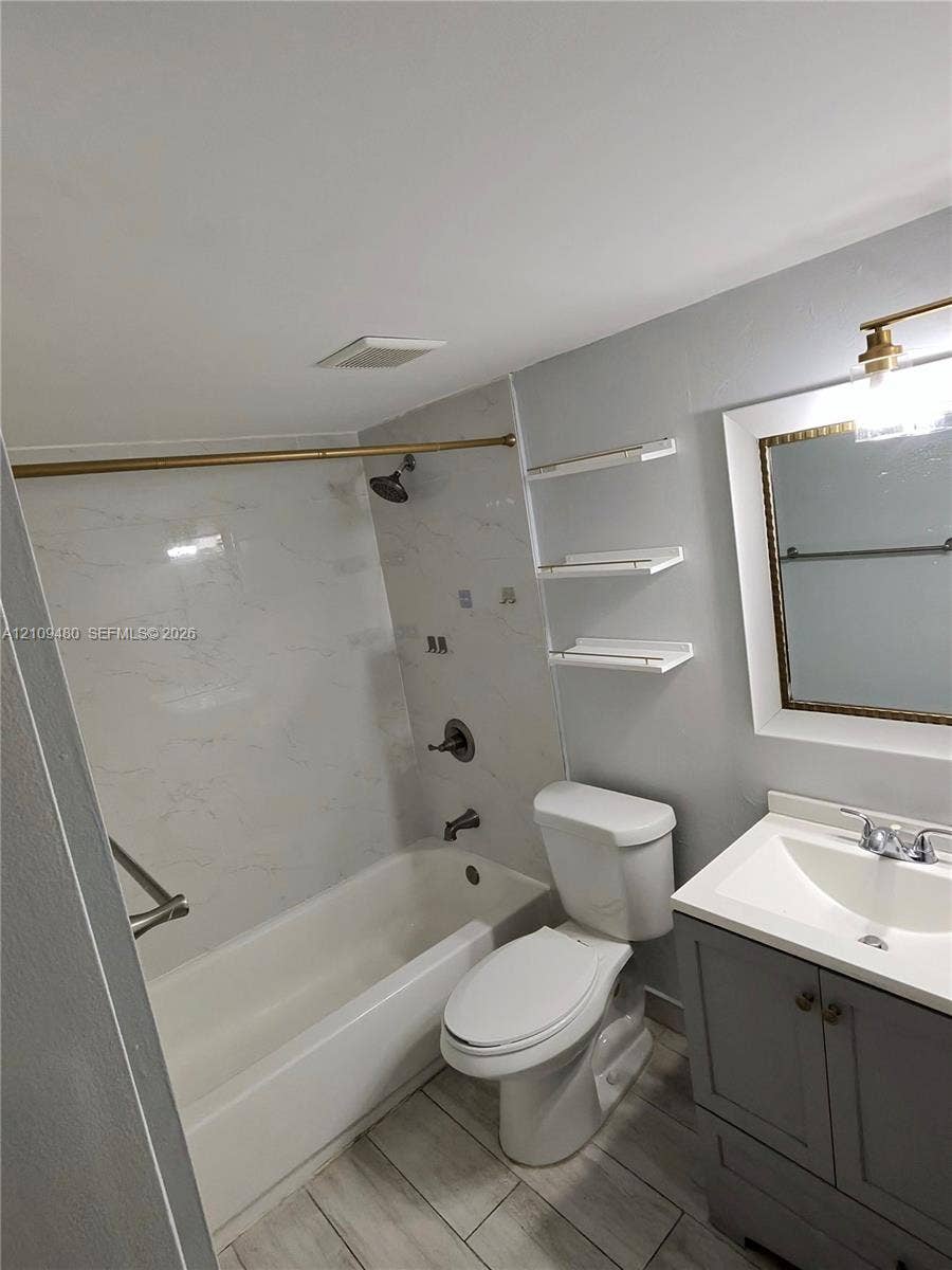 Bathroom featuring a gray vanity with an integrated sink, stone-finish tub surround, wood-finish flooring, wall-mounted shelving, and a framed mirror with a light fixture