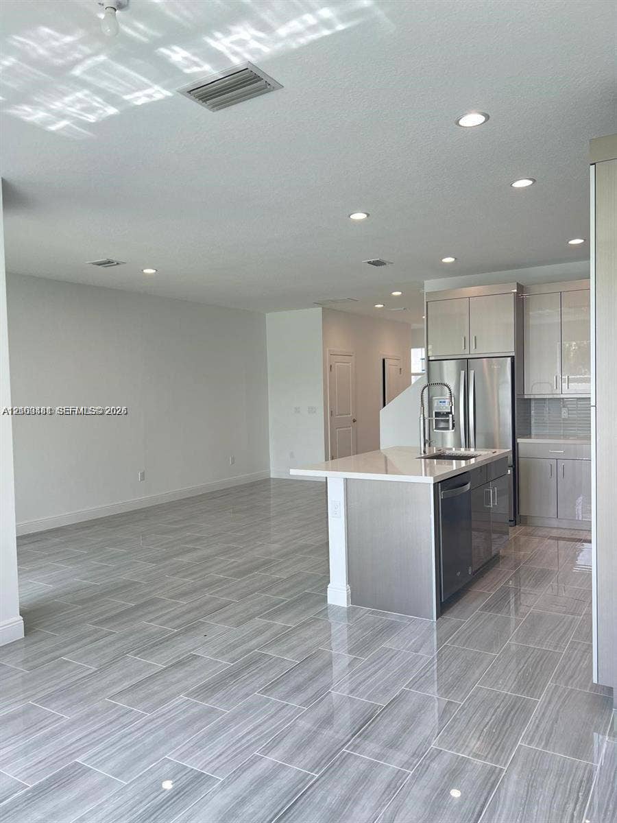 Open-concept kitchen featuring gray-toned cabinetry, high-gloss stone-surface countertops, an island with a stainless steel undermount sink, and polished tile flooring