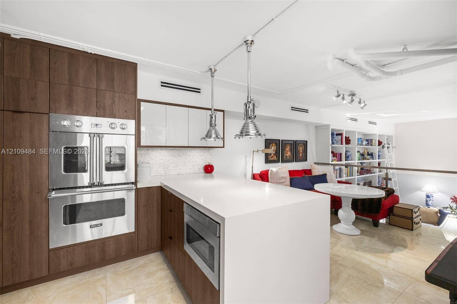 Kitchen featuring wood-finish cabinetry, a waterfall-edge island with stone countertop, stainless steel double wall oven, polished pendant lighting, and built-in floor-to-ceiling shelving