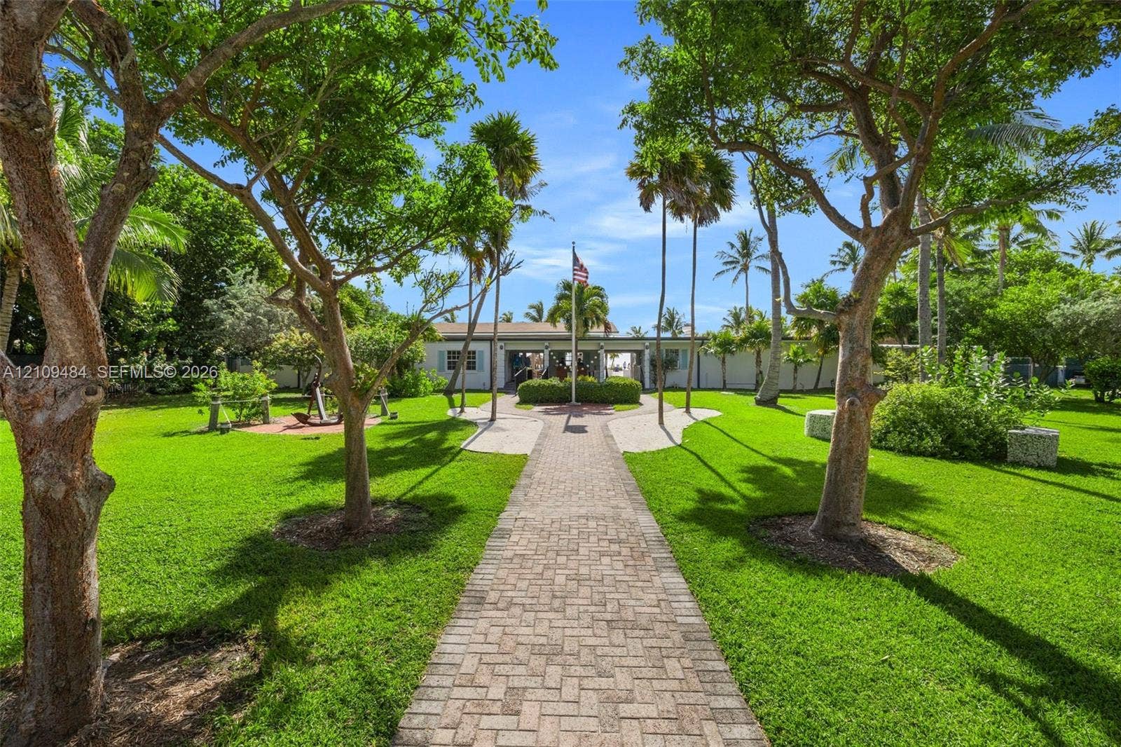 Expansive grounds featuring a brick paver walkway, manicured lawn, established shade trees, and mature tropical landscaping