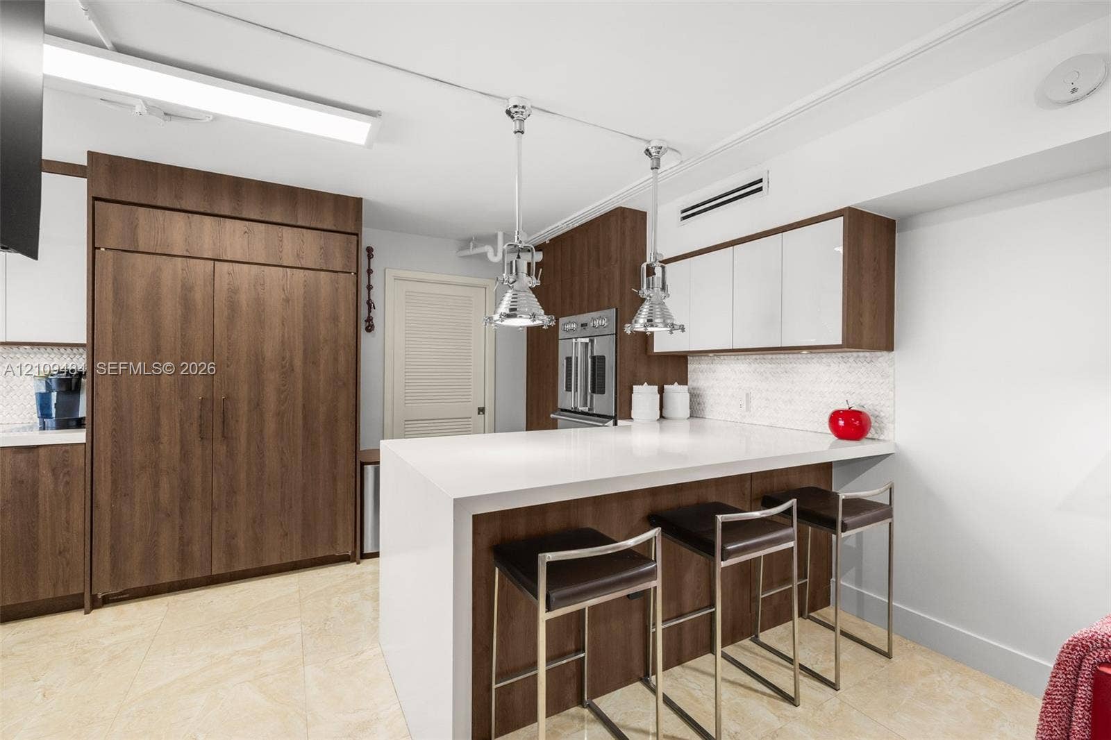 Kitchen featuring a waterfall-edge stone countertop, custom cabinetry with wood-finish and white high-gloss doors, stainless steel wall-mounted oven, and dual polished-metal pendant light fixtures