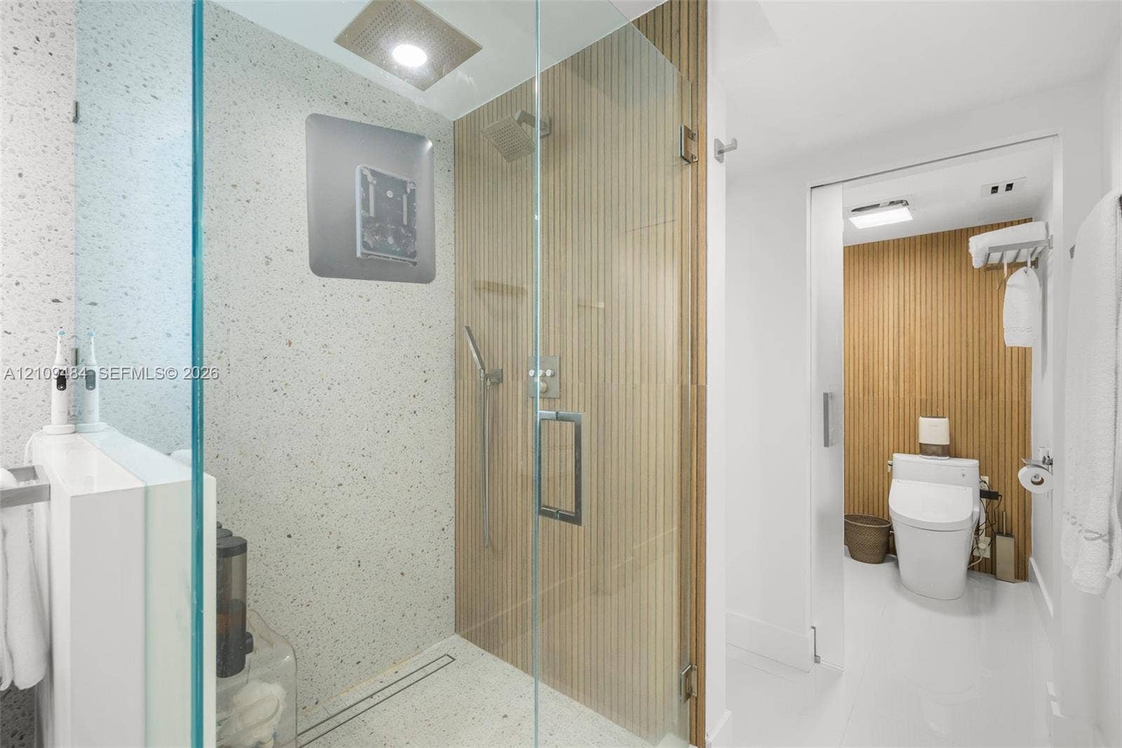 Walk-in shower with terrazzo-style wall and floor tiles, wood-slat accent wall, frameless glass enclosure, rain showerhead, and sliding door entry