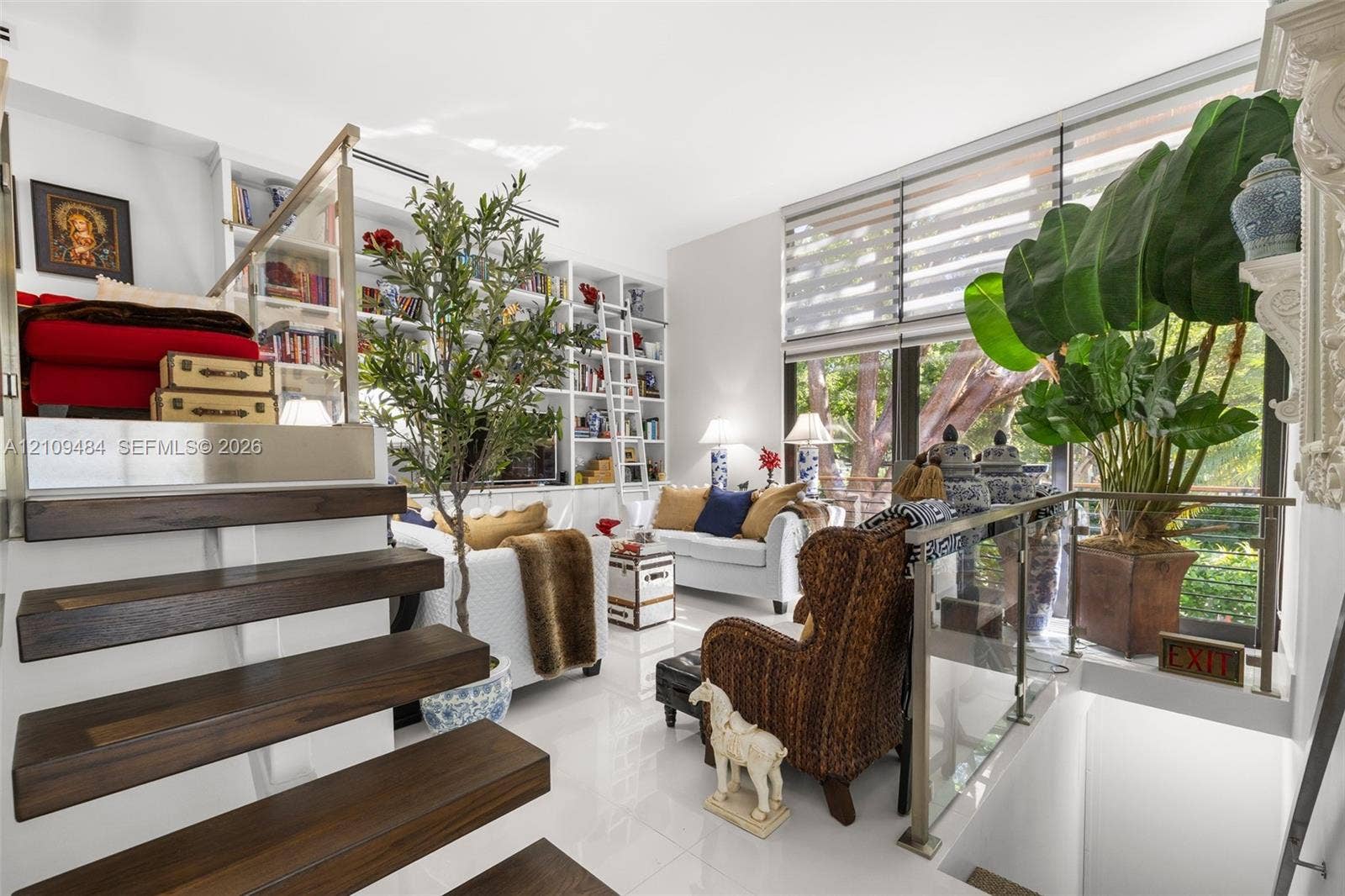 Floor-to-ceiling built-in library shelving with rolling ladder, high-gloss tile flooring, dark wood-finish open staircase, floor-to-ceiling windows with integrated roller shades, and stainless steel and glass stair railings