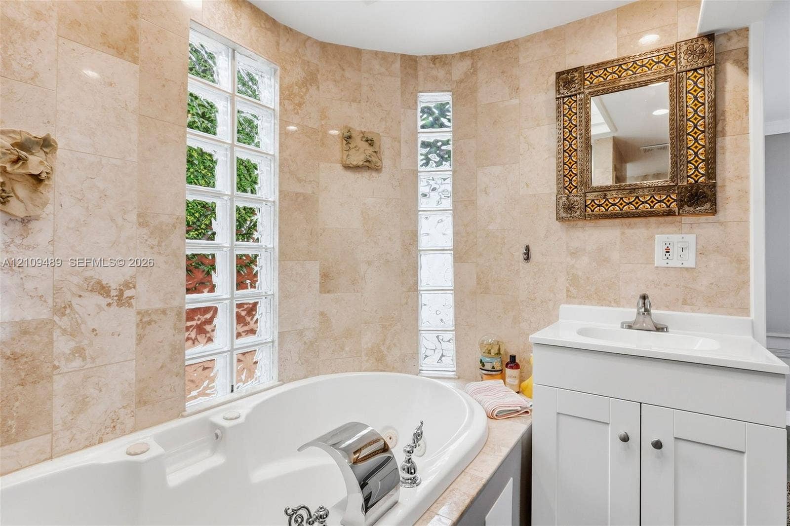 Fort Lauderdale, FL 33312 Curved bathtub surround with stone-tiled walls, glass block windows, built-in soaking tub, chrome waterfall faucet, and white vanity with solid-surface countertop
