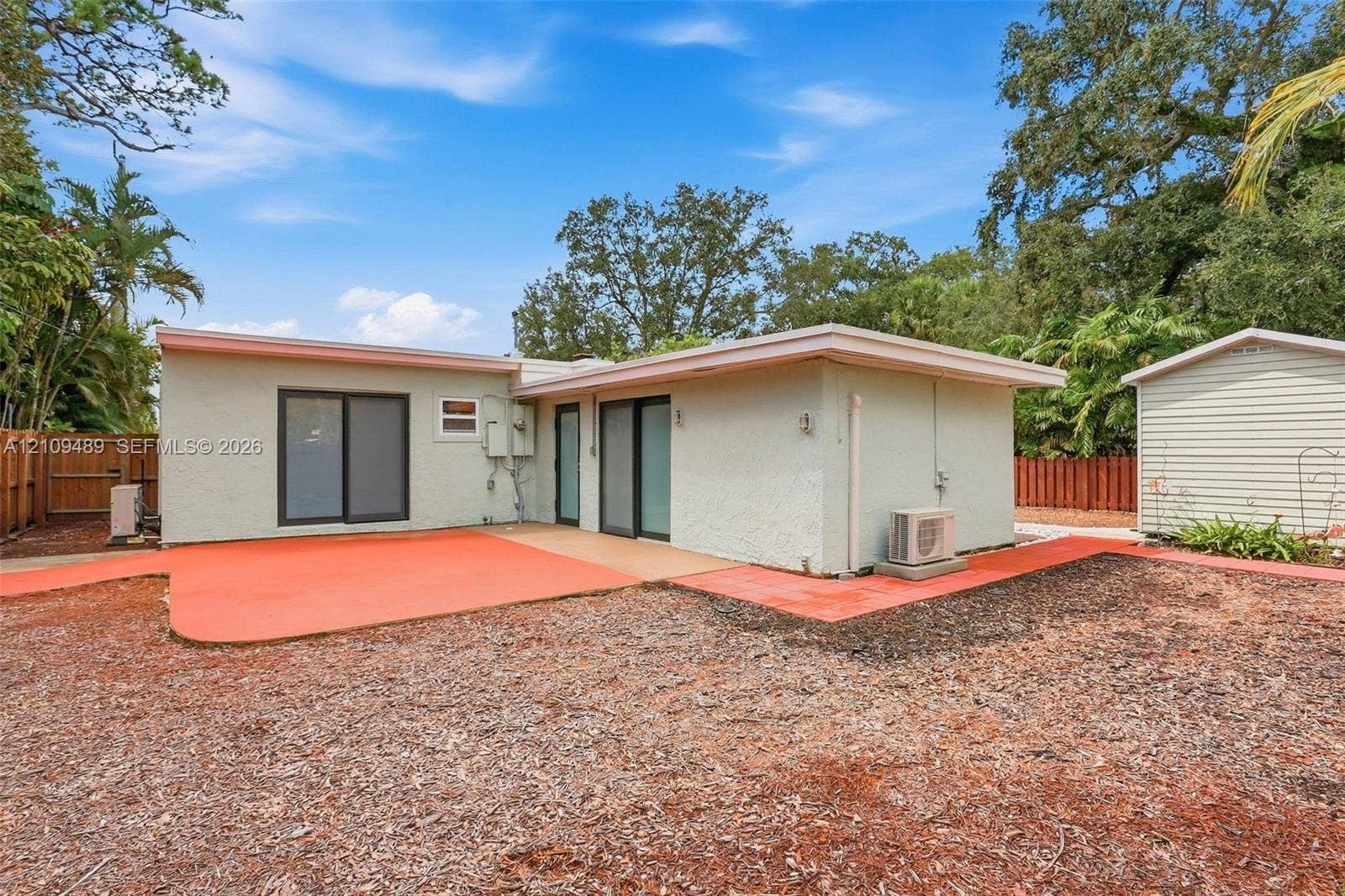 Fort Lauderdale, FL 33312 Stucco exterior featuring dual sliding glass doors, a painted concrete patio, and a detached storage structure enclosed by perimeter wood fencing