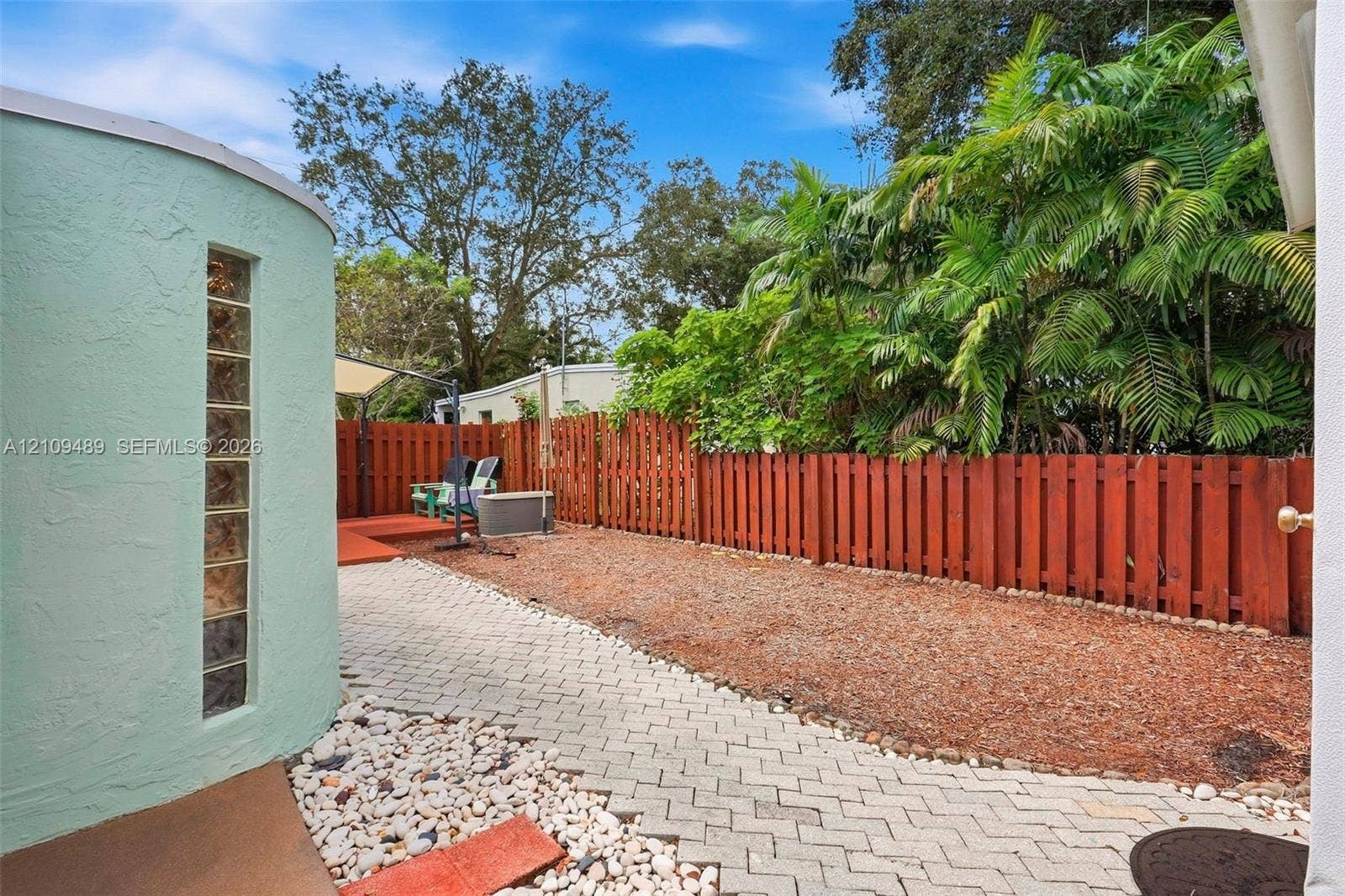 Fort Lauderdale, FL 33312 Backyard featuring a curved exterior wall with a vertical glass block window, stone paver pathway, wood-style privacy fencing, and mature palm trees