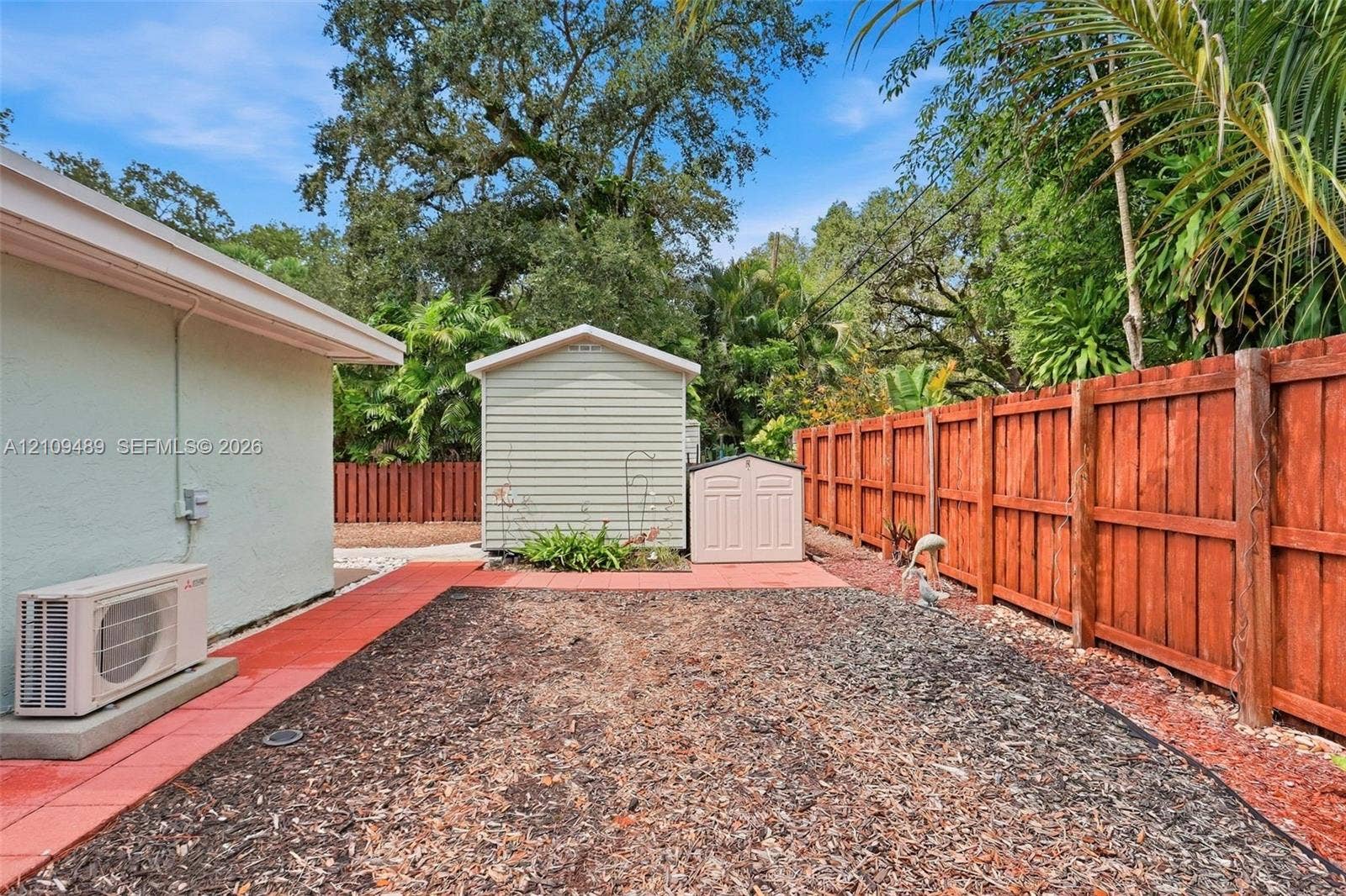 Fort Lauderdale, FL 33312 Rear exterior featuring a wood-style privacy fence, red paver pathways, dedicated storage structures, and mature tree canopy