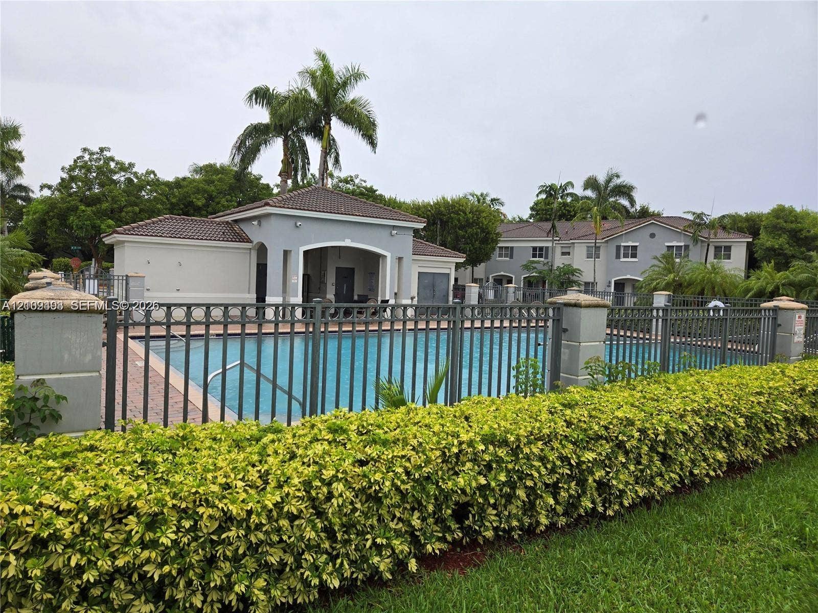 Community pool featuring a concrete deck, metal perimeter fencing, tiled roof clubhouse structure, and manicured landscaping