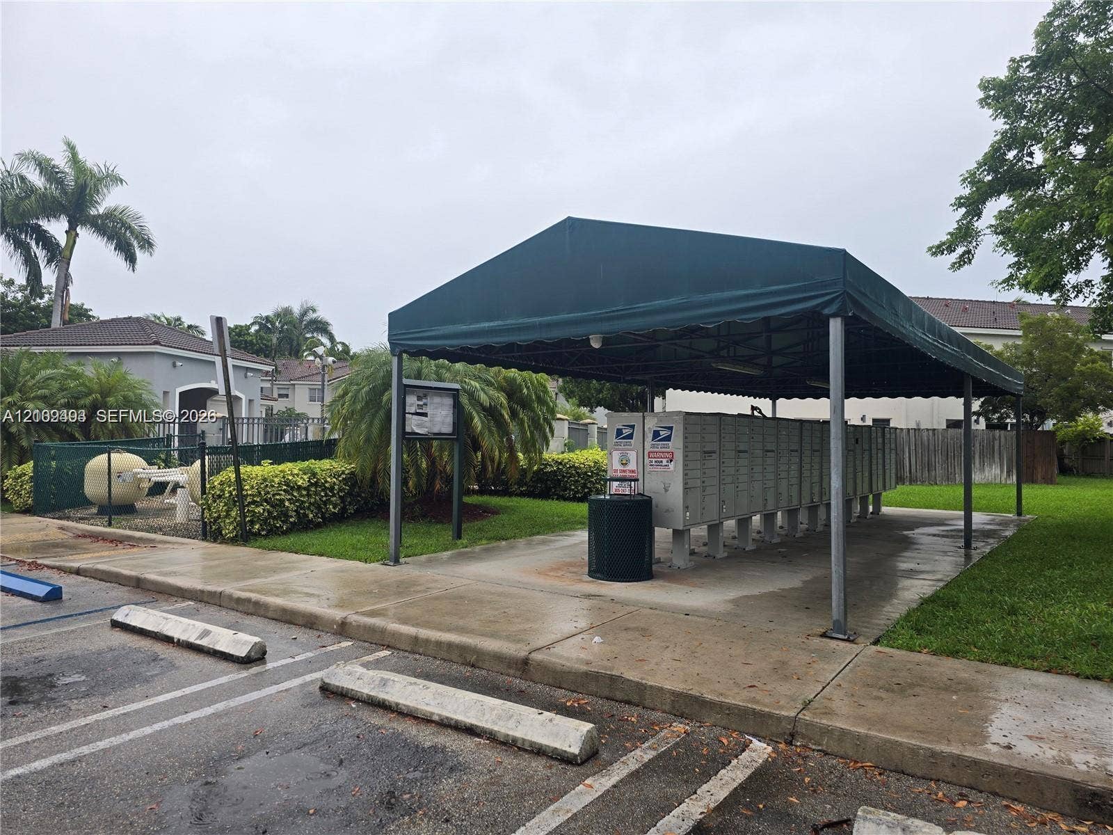 Covered mail kiosk station featuring a dark green canopy, multiple locking postal compartments, and concrete hardscaping