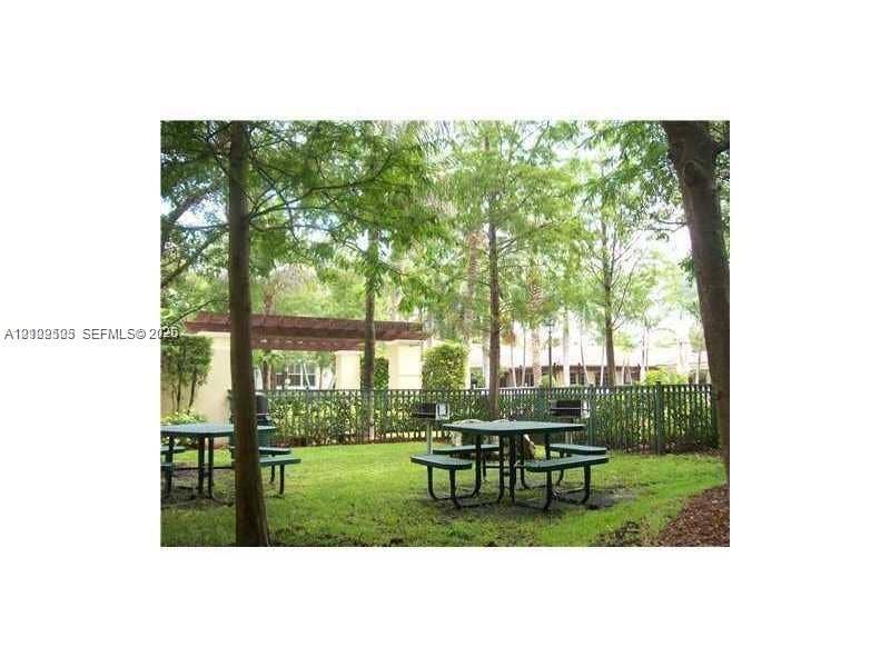 6804 W Sample Road, #6804, Coral Springs, Coral Springs, FL 33067 Outdoor recreation area featuring a wooden pergola, built-in metal grills, and mature trees situated on a grass lawn enclosed by a metal fence