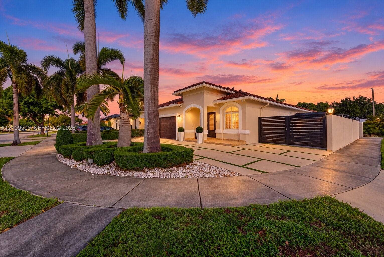 7721 NW 201st Terrace, Hialeah, FL 33015 Stucco exterior featuring an arched entryway, segmented concrete driveway with grass inlays, and gated side perimeter wall, complemented by mature palm trees and manicured landscape hedging