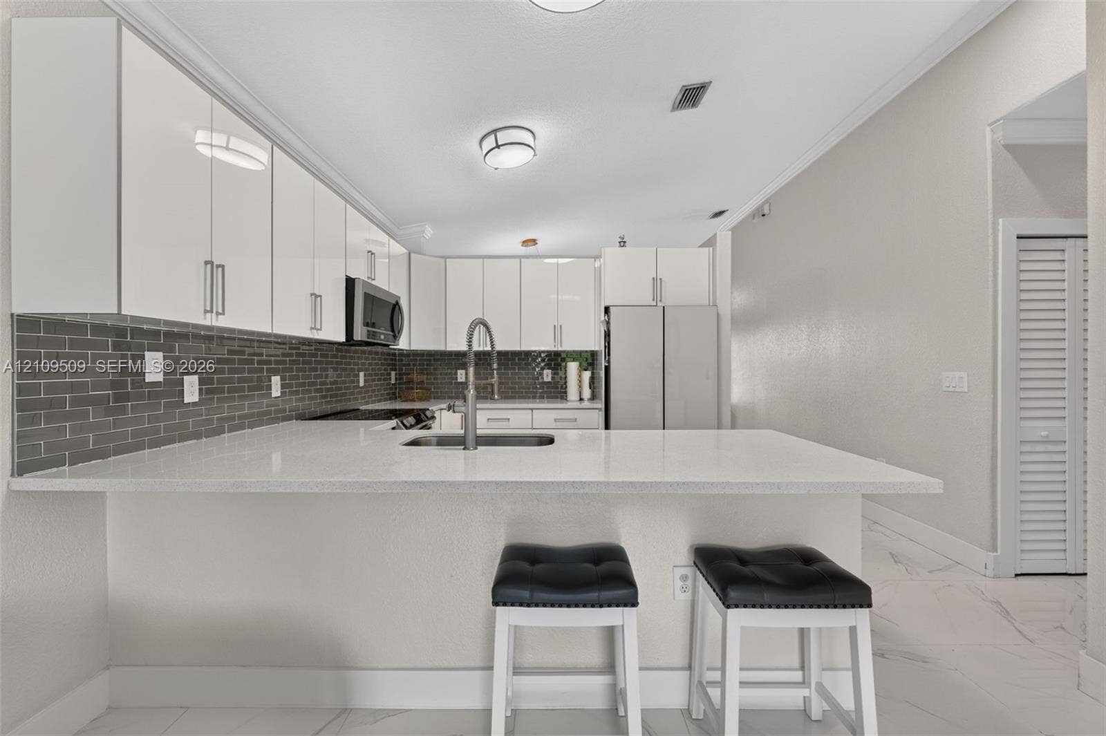 7721 NW 201st Terrace, Hialeah, FL 33015 Kitchen featuring high-gloss white cabinetry, white stone-finish countertops, stainless steel appliances, dark grey subway tile backsplash, and light-toned tile flooring