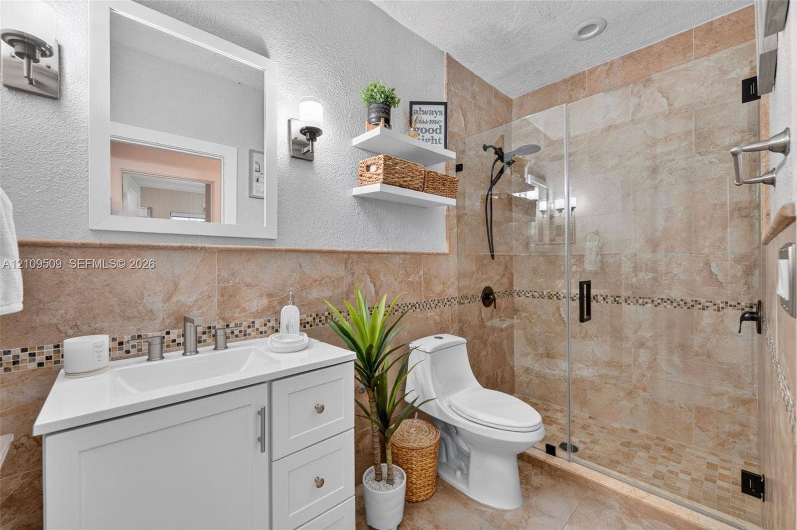 7721 NW 201st Terrace, Hialeah, FL 33015 Bathroom featuring a white vanity with an integrated sink, a glass-enclosed walk-in shower with mosaic tile accents, wall-mounted lighting, and neutral stone-finish wall and floor tiling