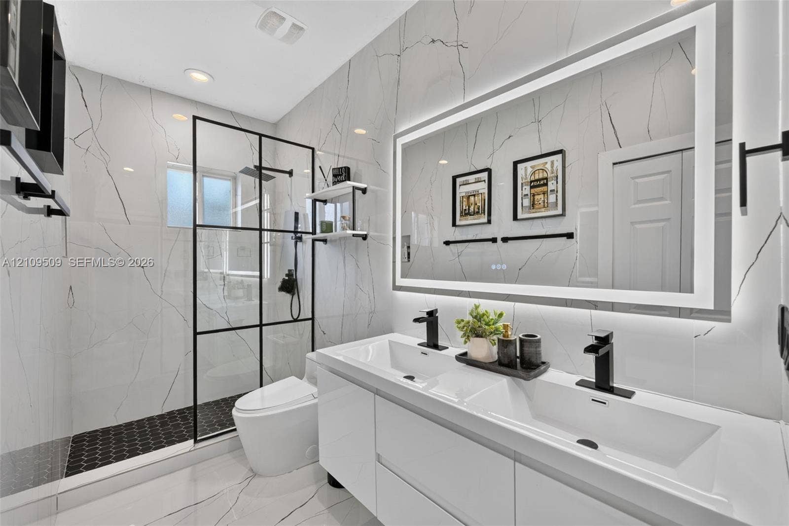 7721 NW 201st Terrace, Hialeah, FL 33015 Modern bathroom featuring large-format veined stone tile walls, a dual-sink vanity with matte black faucets, a frameless glass-enclosed shower with black metal grid framing, and a large backlit vanity mirror