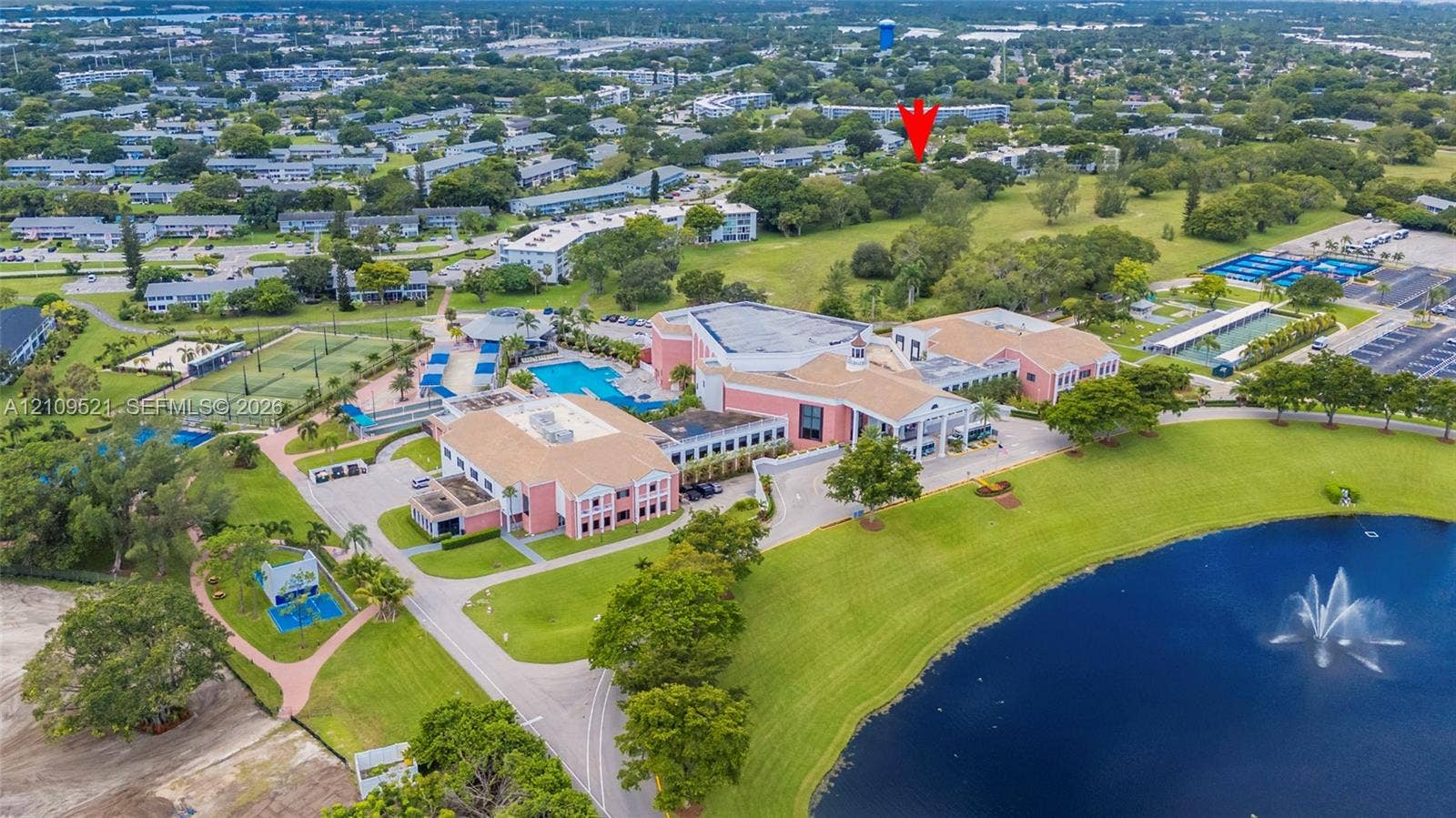 Aerial perspective featuring a multi-building complex, communal swimming pool, tennis courts, and a central water feature with a fountain