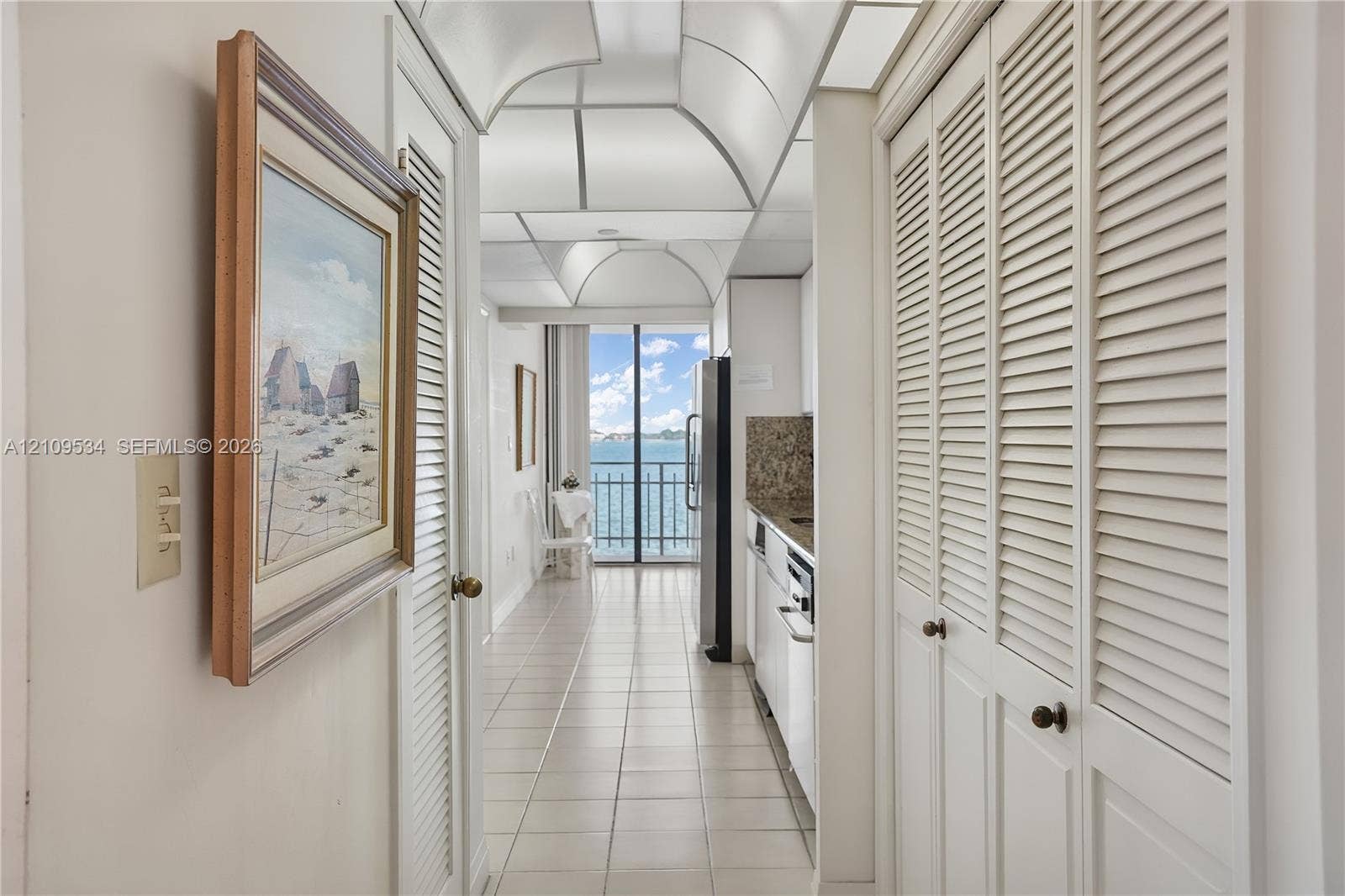 Hallway featuring white tile flooring, louvered closet doors, and a vaulted ceiling, leading to a balcony with water scenery