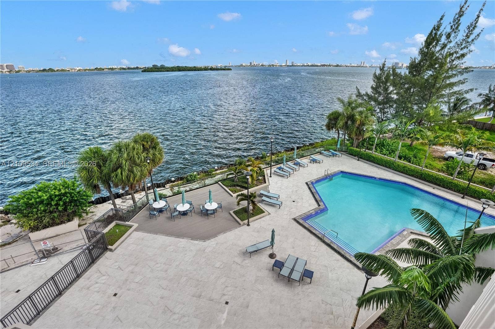Waterfront swimming pool with stone-tiled decking, composite wood-finish platform, metal perimeter fencing, and mature palm trees