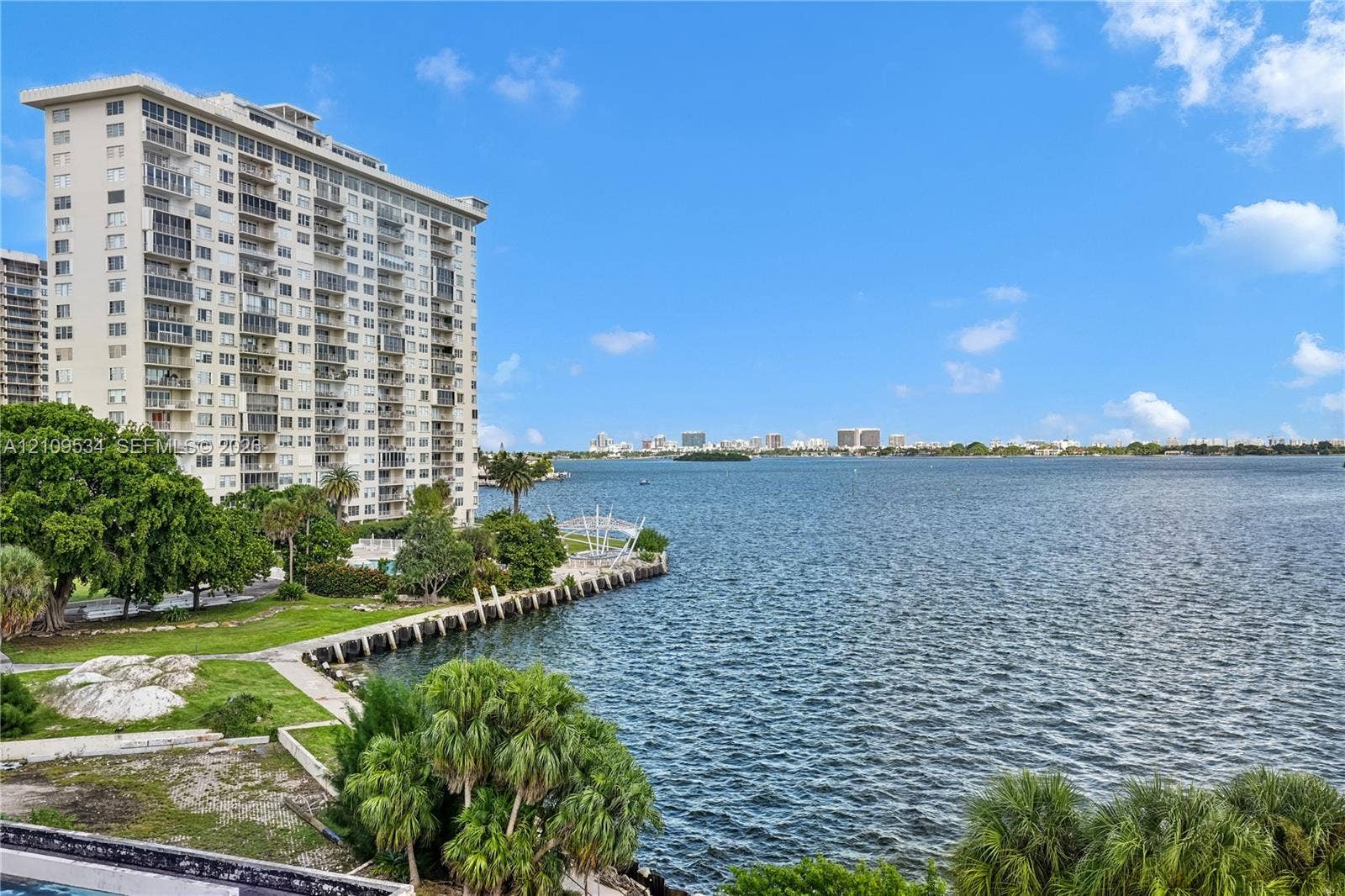High-rise residential exterior with water frontage, concrete bulkhead, mature landscaping, and palm trees