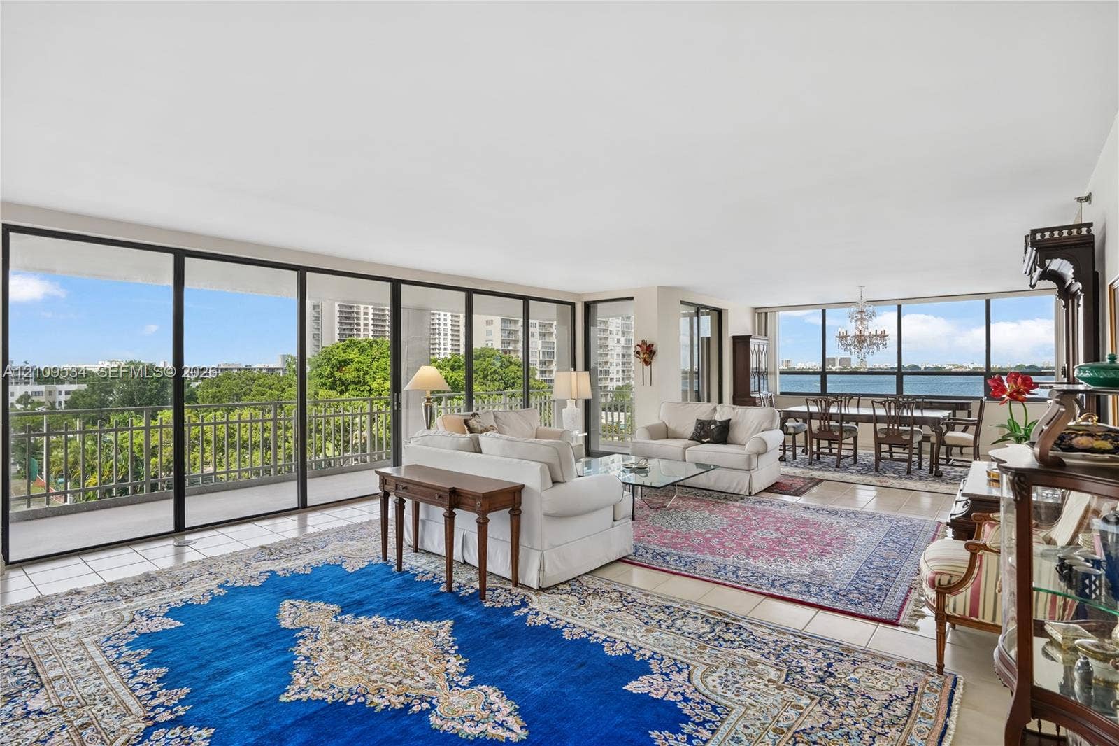 Expansive living area featuring floor-to-ceiling windows, tile flooring, a balcony with metal railing, and decorative chandelier lighting