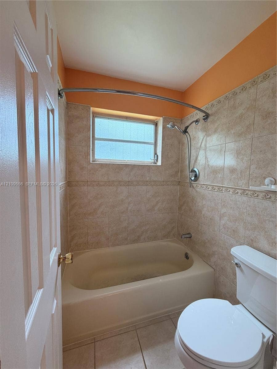 Bathtub with a curved shower rod, wall-mounted hand-held shower fixture, neutral tile surround with decorative inlay, and tile flooring