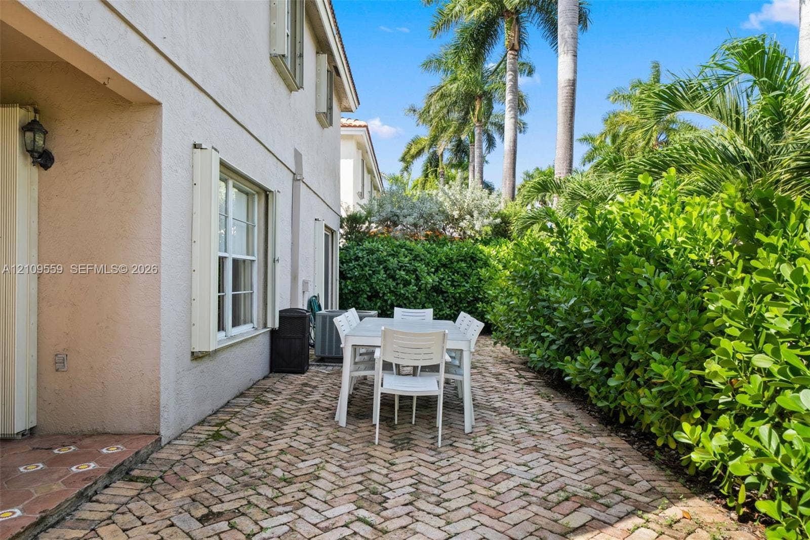 10217 NW 56th Street, Doral, FL 33178 Herringbone-patterned brick paver patio featuring stucco exterior walls, multi-pane windows, and manicured perimeter hedges