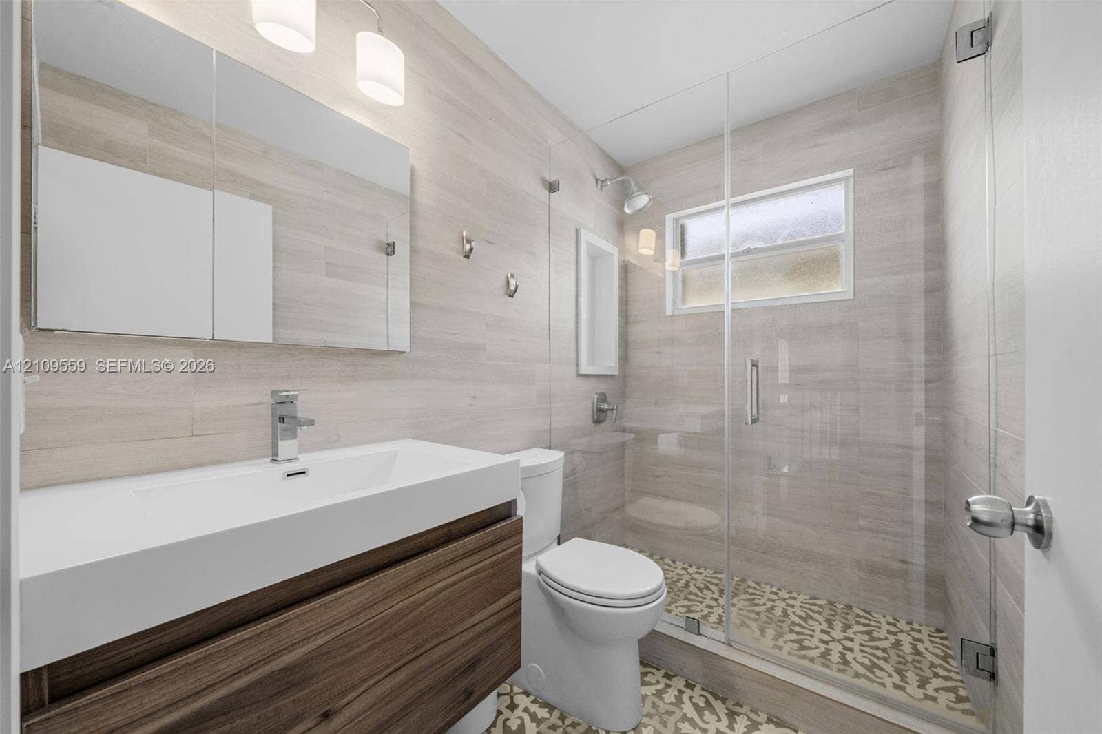 10217 NW 56th Street, Doral, FL 33178 Floating vanity with a rectangular sink, wood-finish cabinetry, frameless glass shower enclosure, patterned tile shower flooring, and light-toned horizontal wall tile