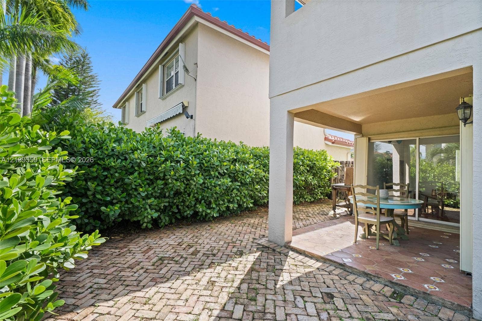 10217 NW 56th Street, Doral, FL 33178 Covered patio with ceramic tile flooring, brick paver hardscaping, mature privacy hedges, and a wall-mounted exterior light fixture