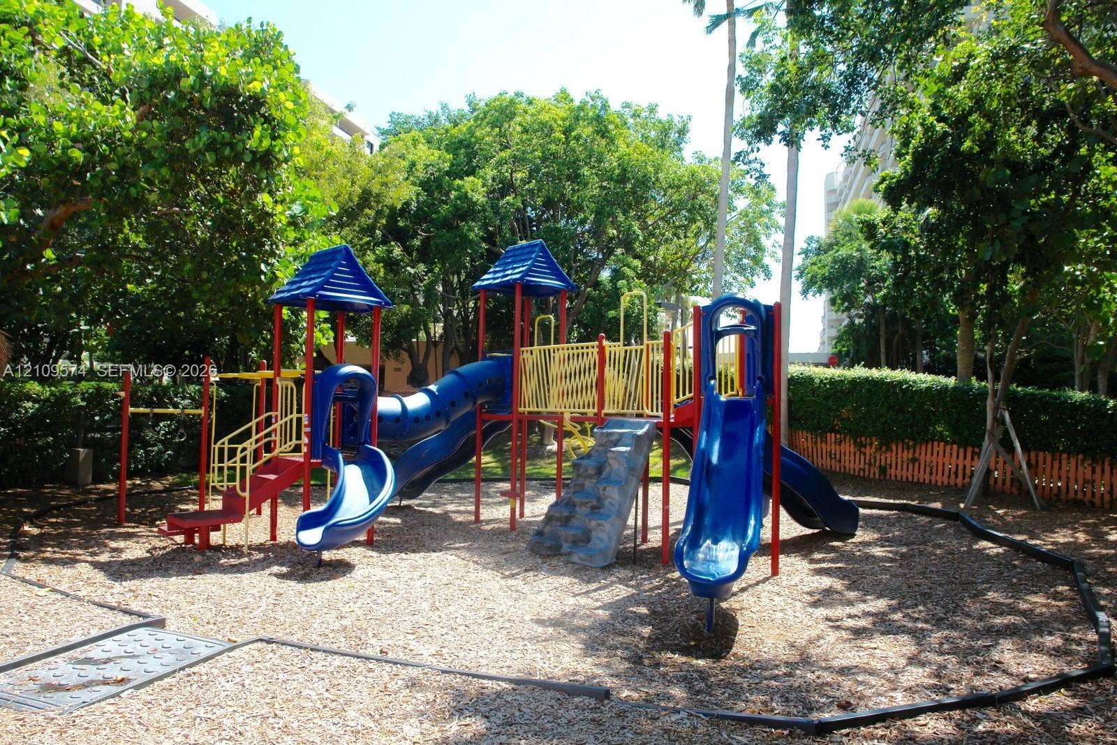 Outdoor recreation area featuring a multi-level playground structure, blue slides, climbing wall, and wood chip ground cover surrounded by mature trees