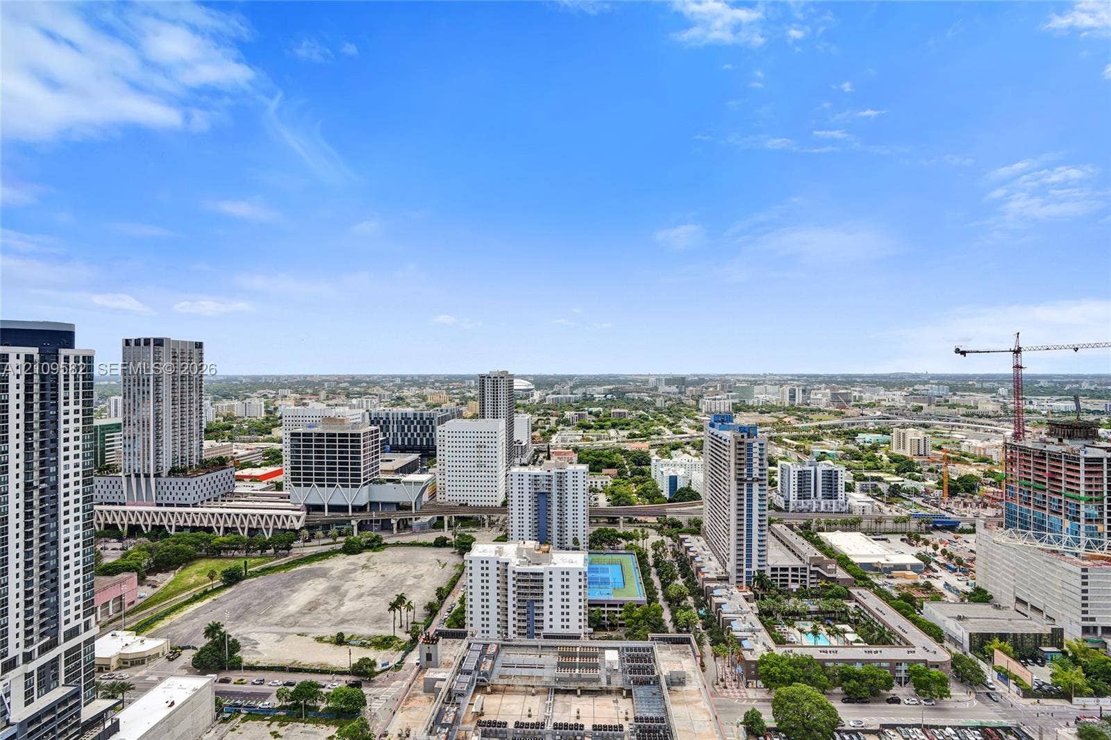 High-rise aerial perspective of an urban skyline featuring prominent skyscrapers, multi-story residential buildings, paved roadways, and a tennis court