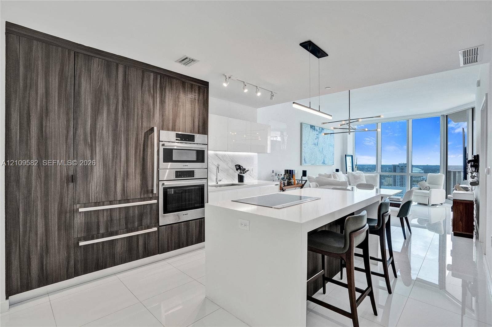Contemporary kitchen featuring white solid-surface countertops, floor-to-ceiling wood-finish cabinetry, stainless steel wall-mounted appliances, and glass floor-to-ceiling windows