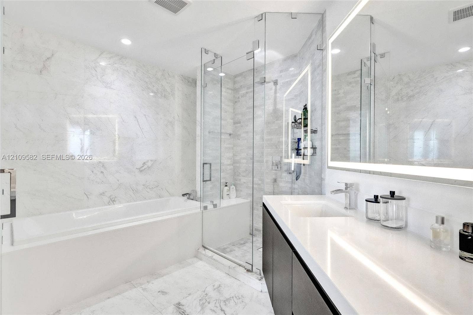 Bathroom featuring a deep soaking tub, glass-enclosed shower with tiled walls, stone-finish flooring, dual-sink vanity with solid-surface countertop, and an integrated LED-lighted mirror