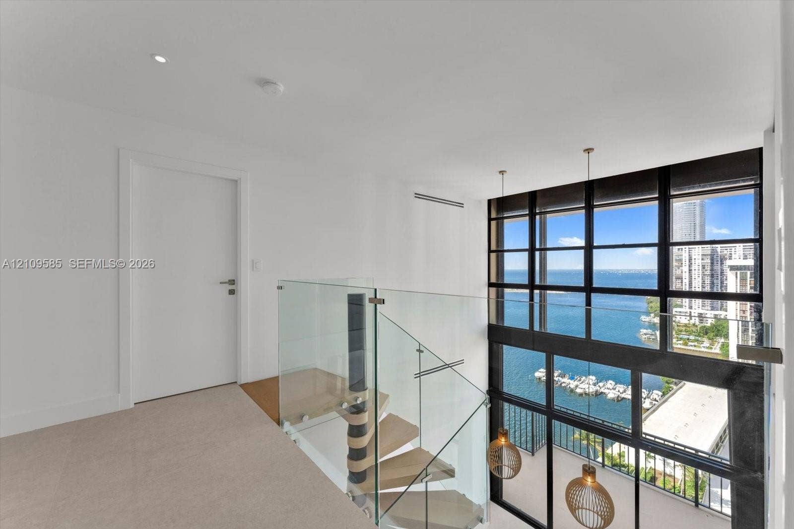 1865 Brickell Avenue, #APHVII, Miami, FL 33129 Double-height floor-to-ceiling windows featuring water frontage, architectural spiral staircase with wood-finish treads, glass panel railing, and integrated pendant lighting