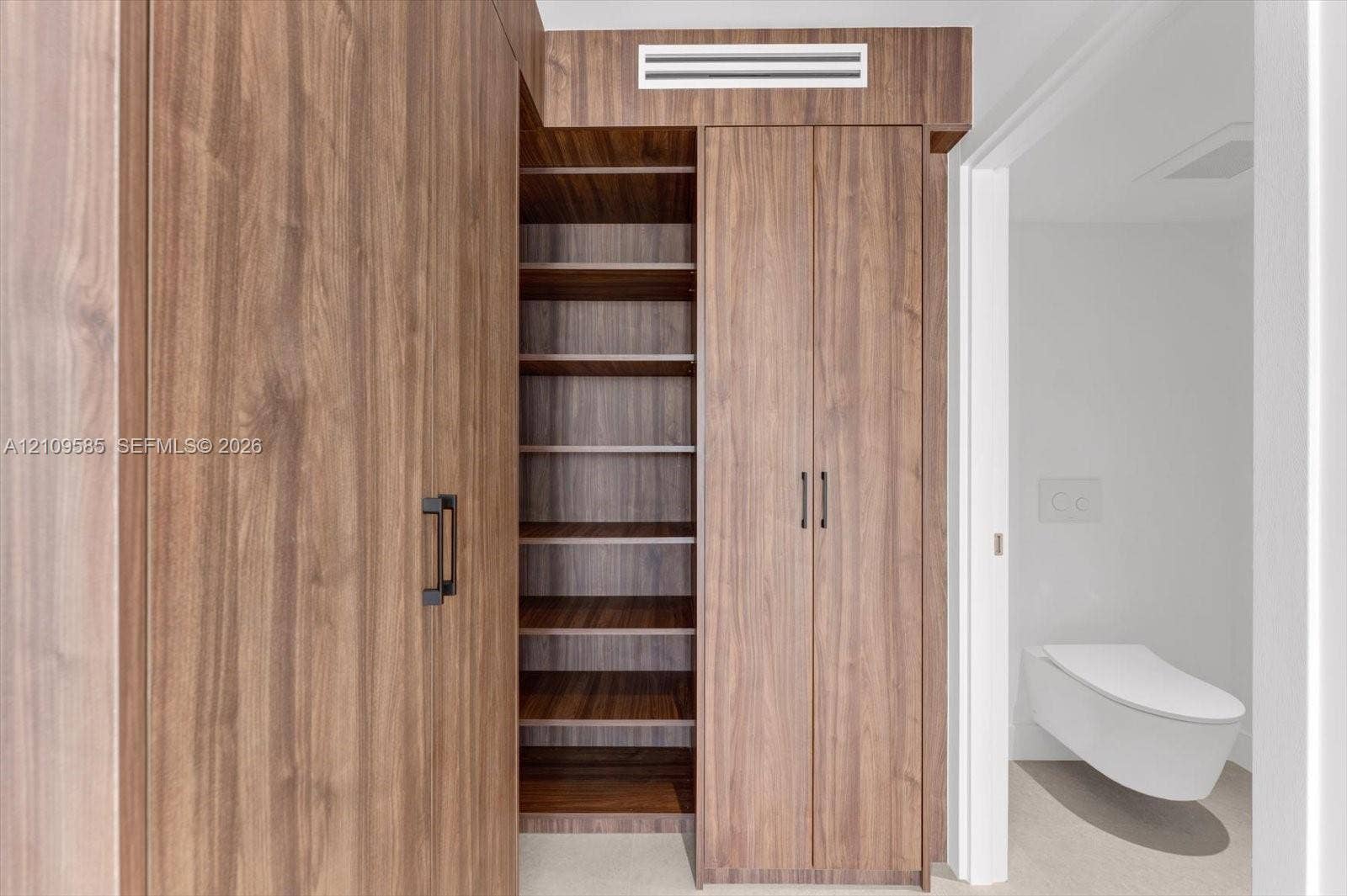 1865 Brickell Avenue, #APHVII, Miami, FL 33129 Floor-to-ceiling wood-finish cabinetry featuring open shelving, double-door storage, matte black hardware, and a wall-mounted toilet