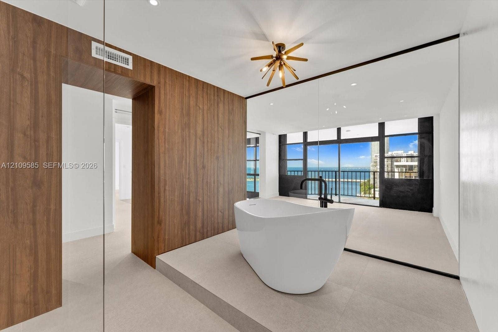 1865 Brickell Avenue, #APHVII, Miami, FL 33129 Freestanding soaking tub with matte black faucet fixture, elevated platform, wood-finish wall paneling, multi-light geometric chandelier, and floor-to-ceiling windows