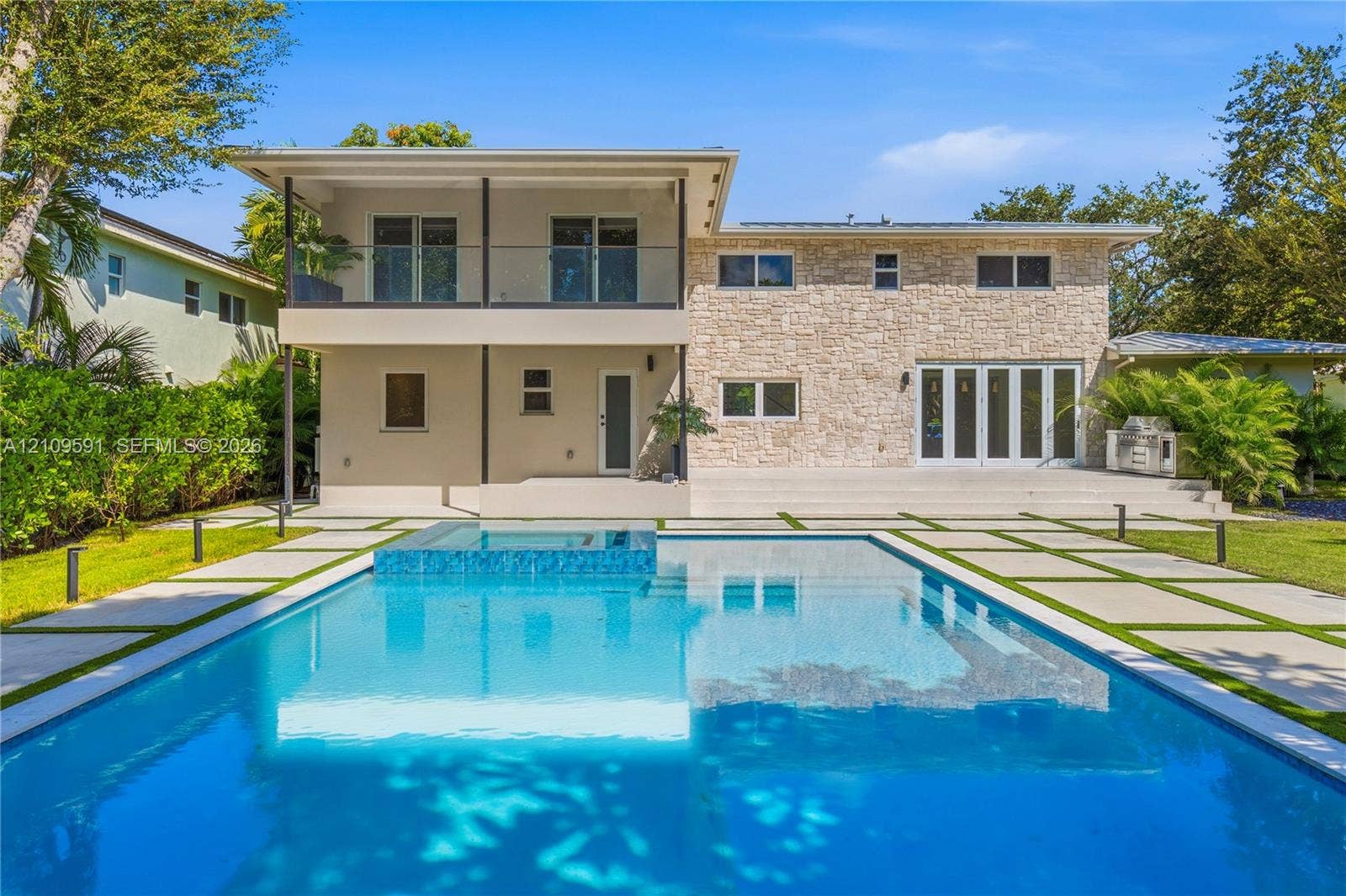 864 NE 91st Terrace, Miami Shores, FL 33138 Exterior facade featuring stone veneer cladding, a second-level balcony with glass railing, a rectangular swimming pool with an integrated spa, and concrete paver hardscaping
