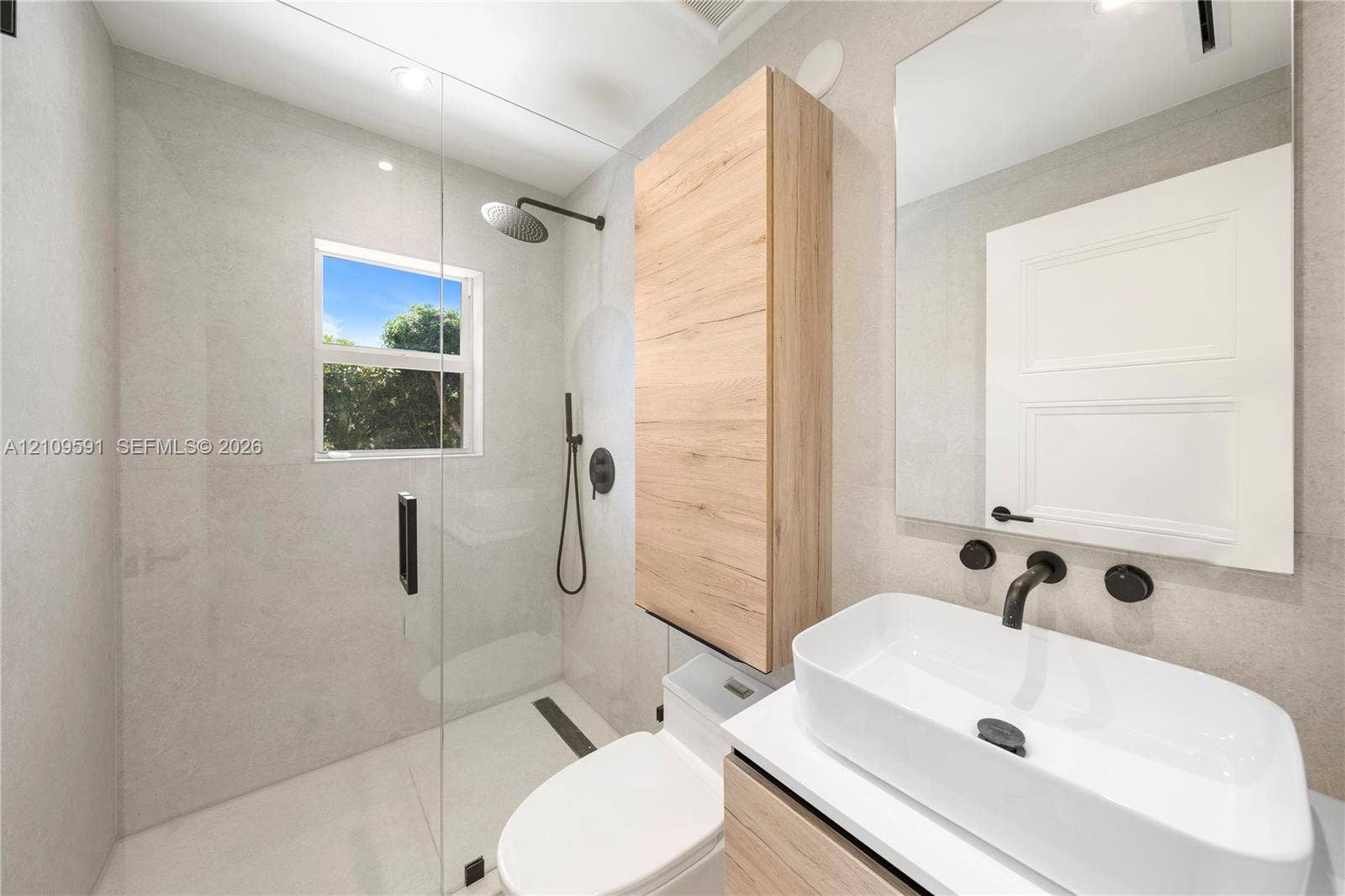 864 NE 91st Terrace, Miami Shores, FL 33138 Walk-in shower with a glass enclosure and wall-mounted matte black rainfall shower head, wood-finish wall-mounted cabinetry, wall-mounted rectangular vessel sink with matte black hardware, and large-format neutral wall tile