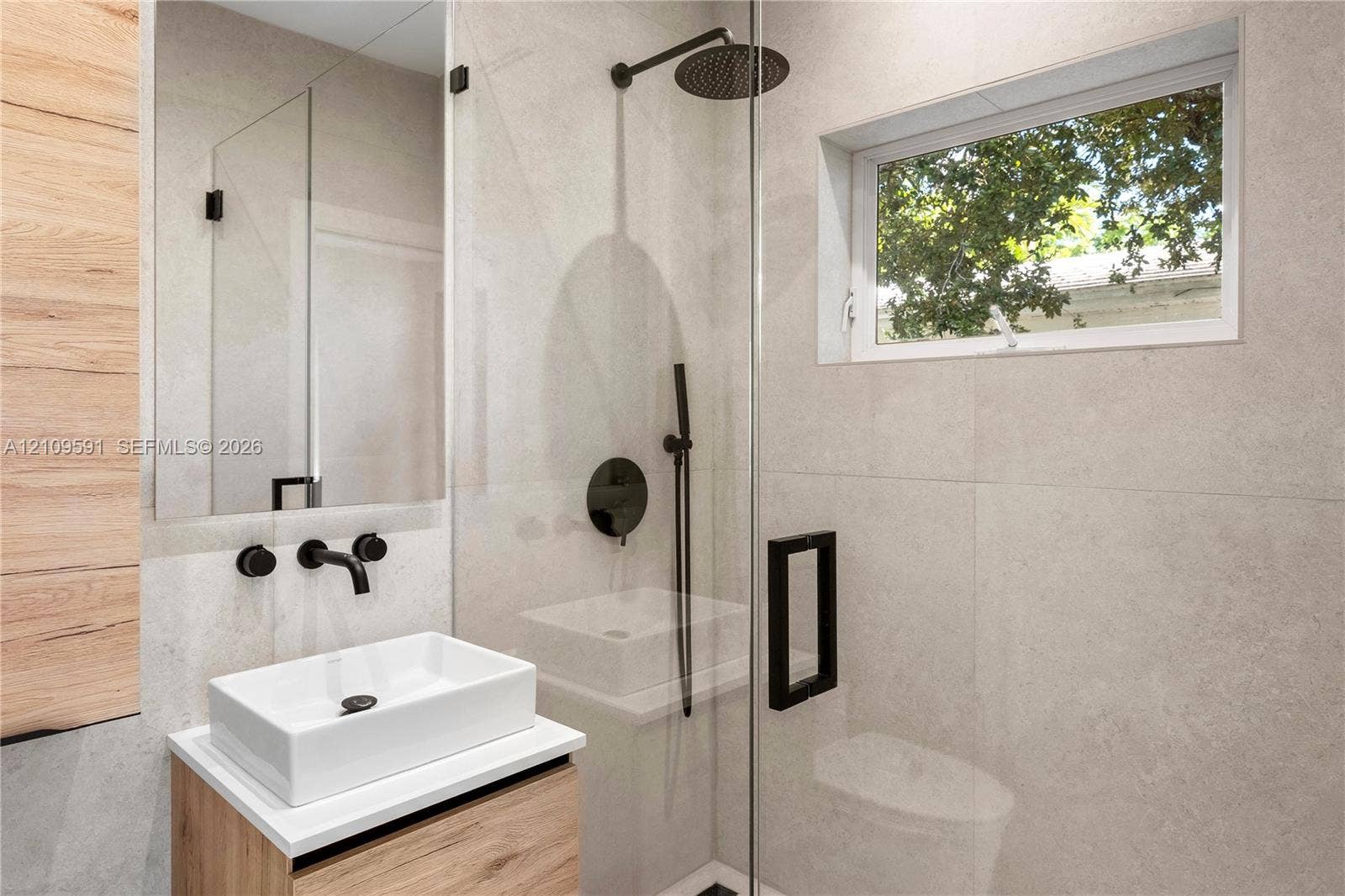 864 NE 91st Terrace, Miami Shores, FL 33138 Modern bathroom featuring large-format stone-finish wall tiles, a glass-enclosed shower with matte black rainfall hardware, a wood-finish floating vanity, and a rectangular vessel sink