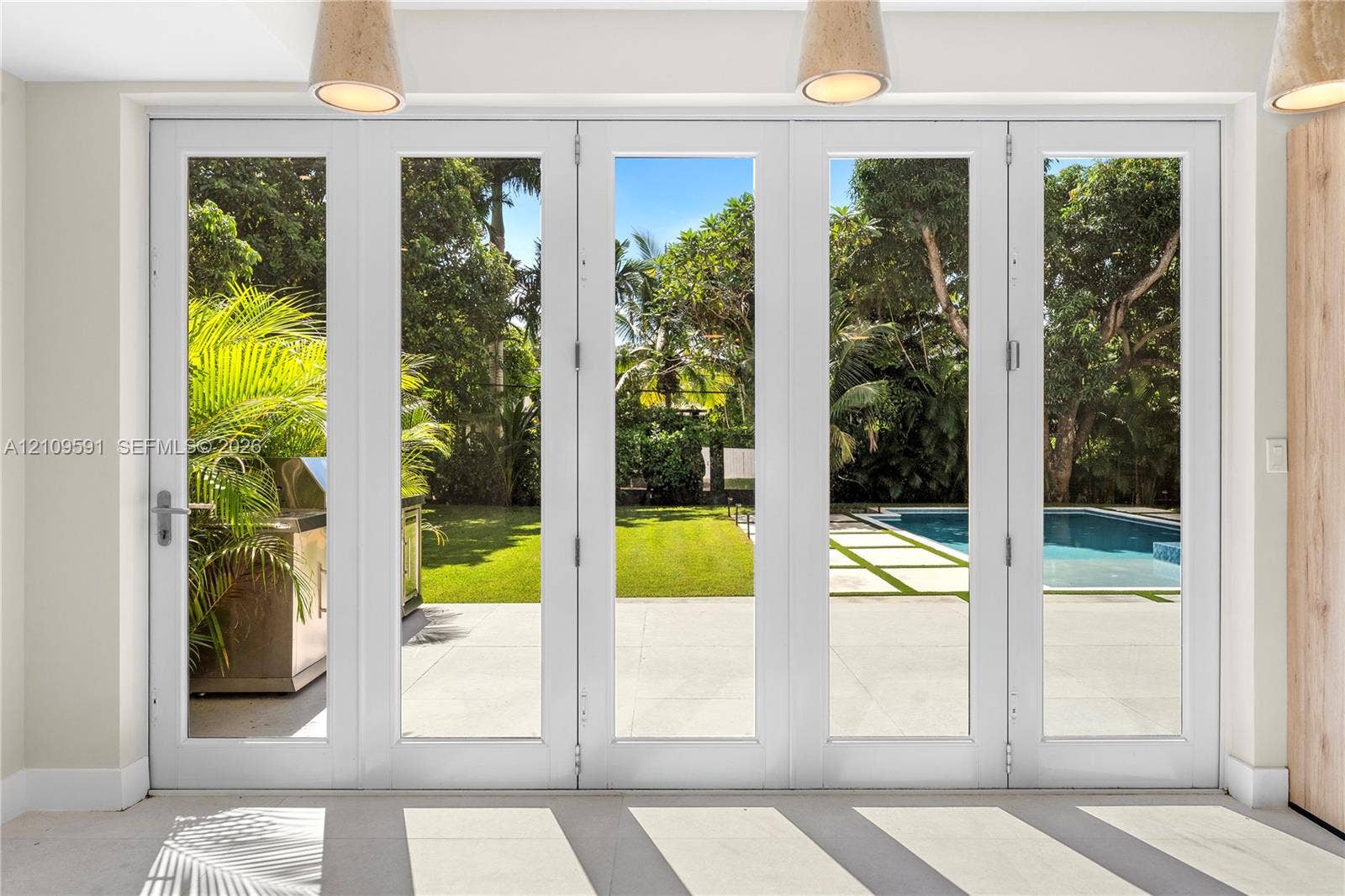 864 NE 91st Terrace, Miami Shores, FL 33138 Multi-panel white-frame glass doors, stone patio, swimming pool, manicured lawn, and suspended pendant lighting
