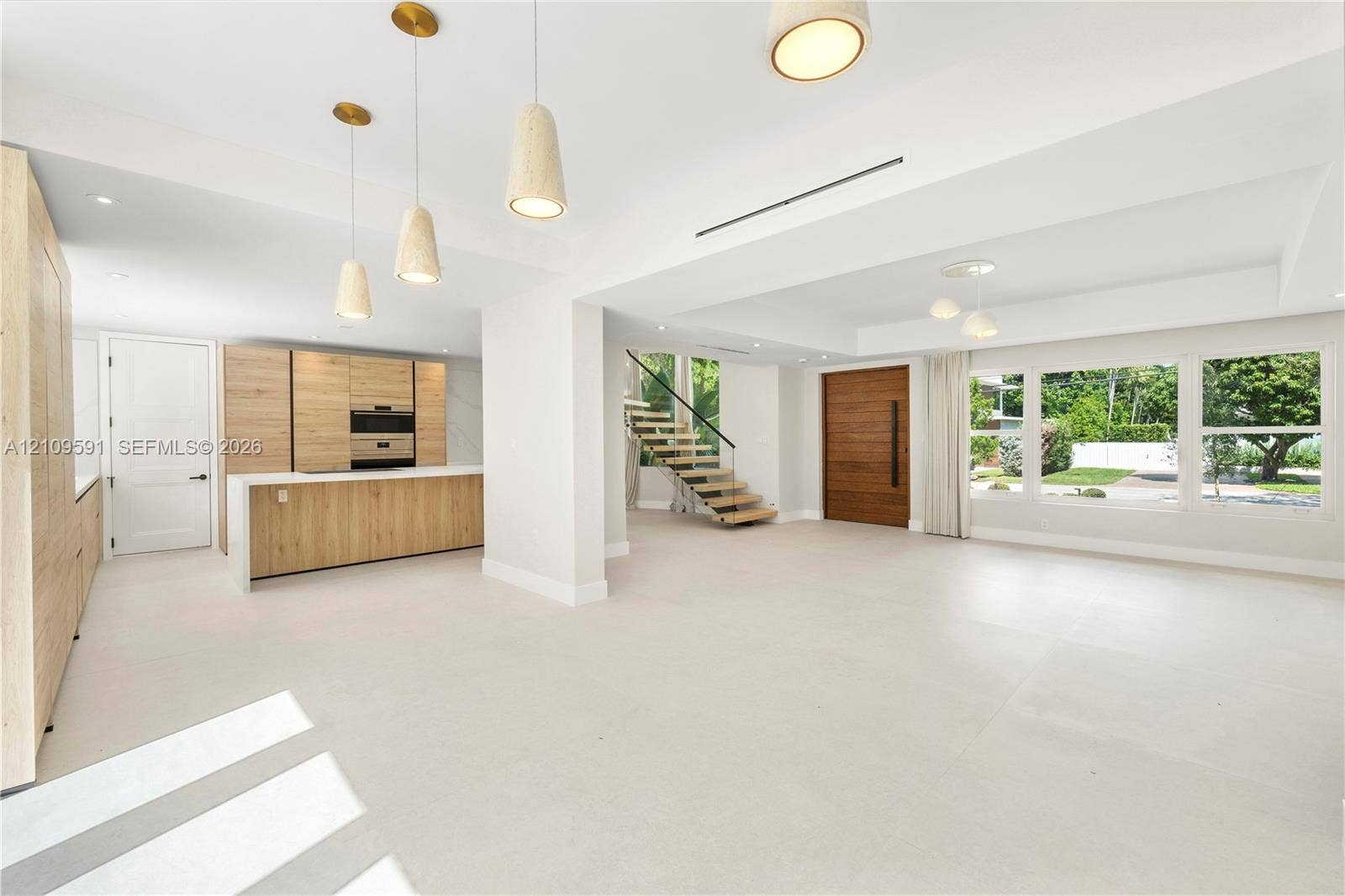864 NE 91st Terrace, Miami Shores, FL 33138 Open-concept interior featuring wood-finish cabinetry, a central kitchen island, large-format tile flooring, a floating staircase with glass railing, and a floor-to-ceiling solid wood entry door